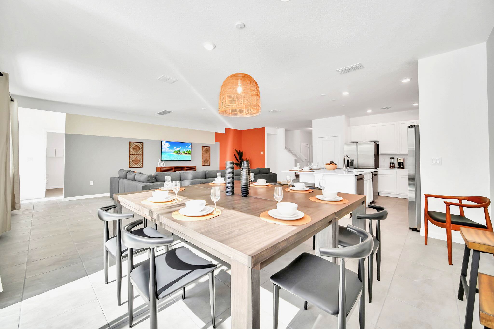Bright dining room featuring a large square wooden table with black chairs and a woven pendant light, offering a view into the modern white kitchen and the living room beyond.