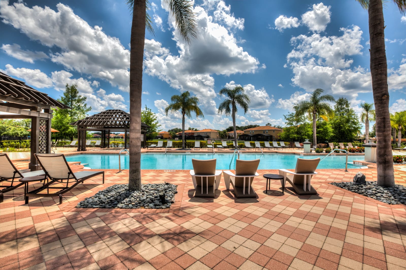 A wide shot of a communal swimming pool area featuring a large blue pool, surrounded by red and tan paver decking, with multiple lounge chairs, tall palm trees, and residential buildings in the distance under a dramatic cloudy sky.