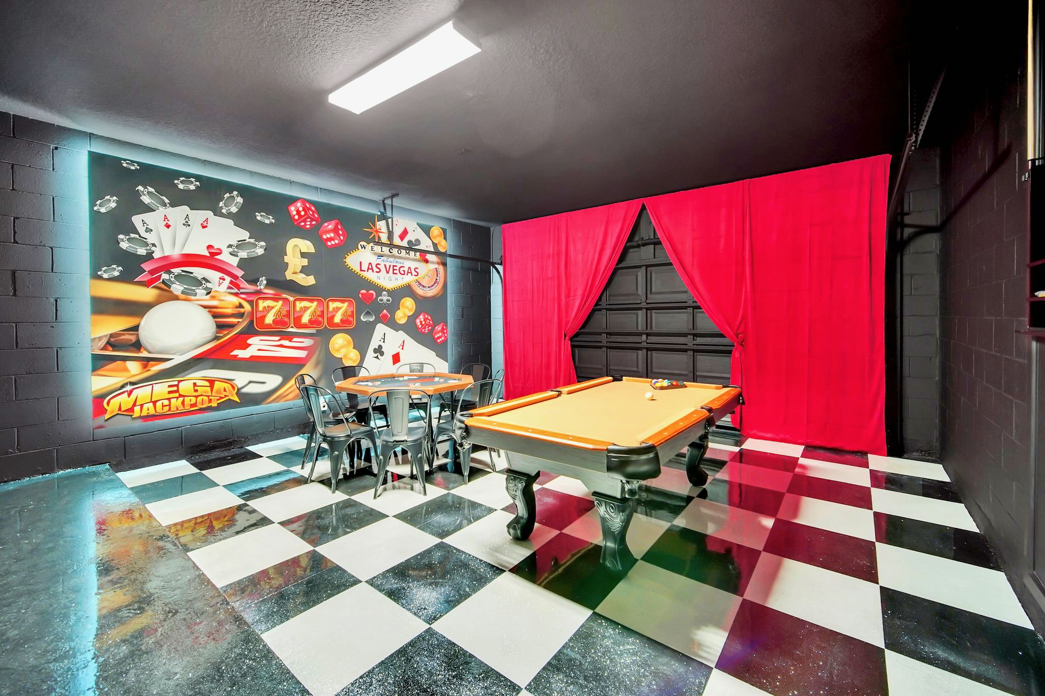 Vibrant game room with a black and white checkered floor, featuring a pool table, a poker table with chairs, and a large, colorful Las Vegas-themed mural on the wall, accented by red curtains.