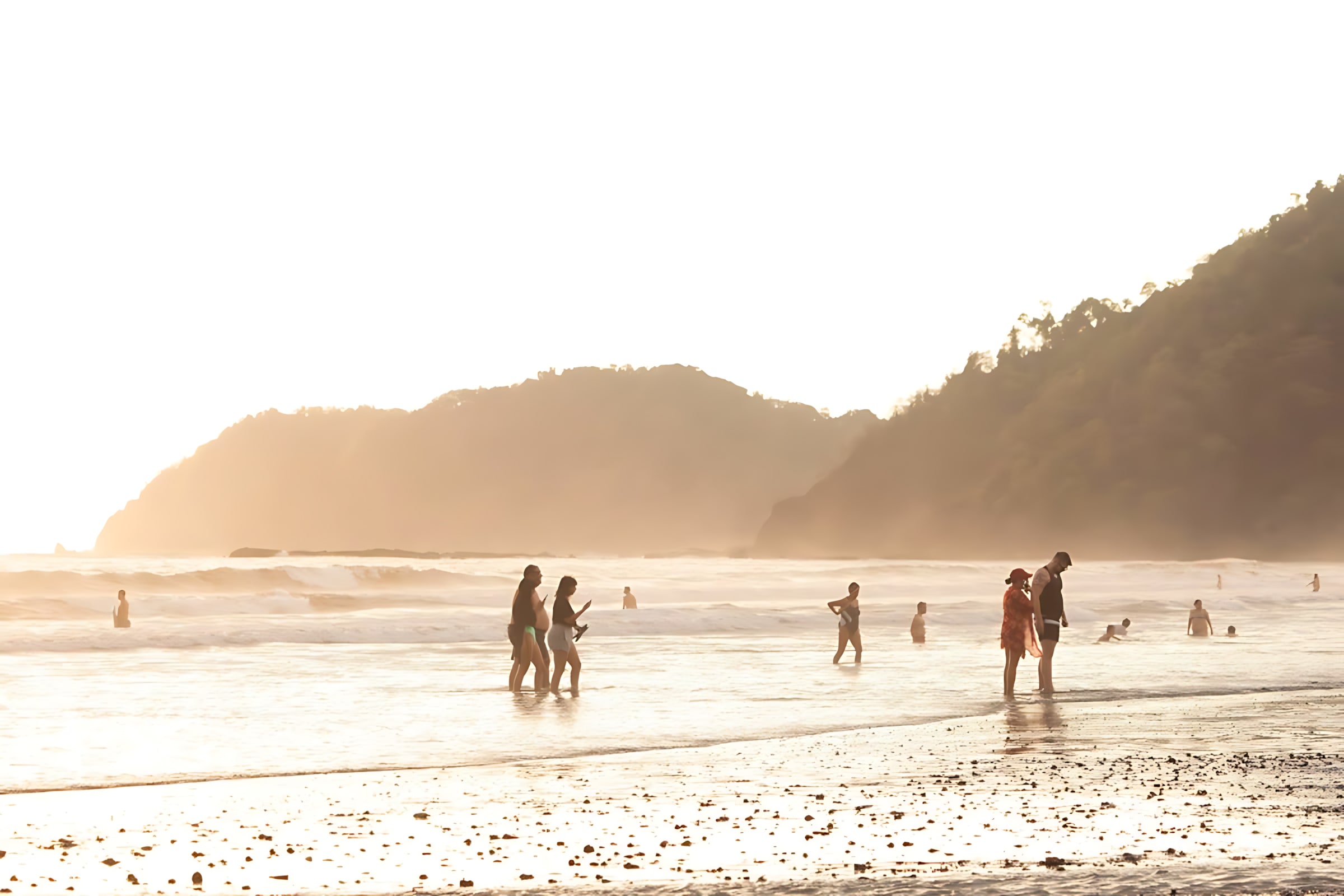 Scenic view of people wading in the shallow ocean water at sunset, with soft golden light illuminating the waves and distant hills.