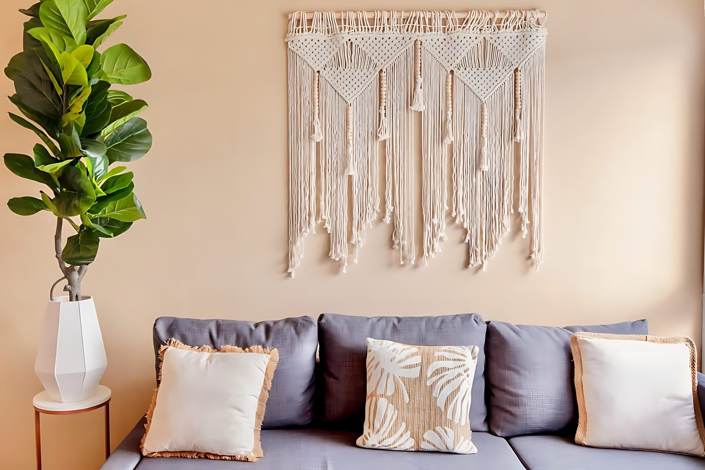 Close-up of the grey sectional sofa with cream and patterned throw pillows, positioned beneath a large, intricate macrame wall hanging. A tall potted plant in a white geometric planter adds greenery to the corner.