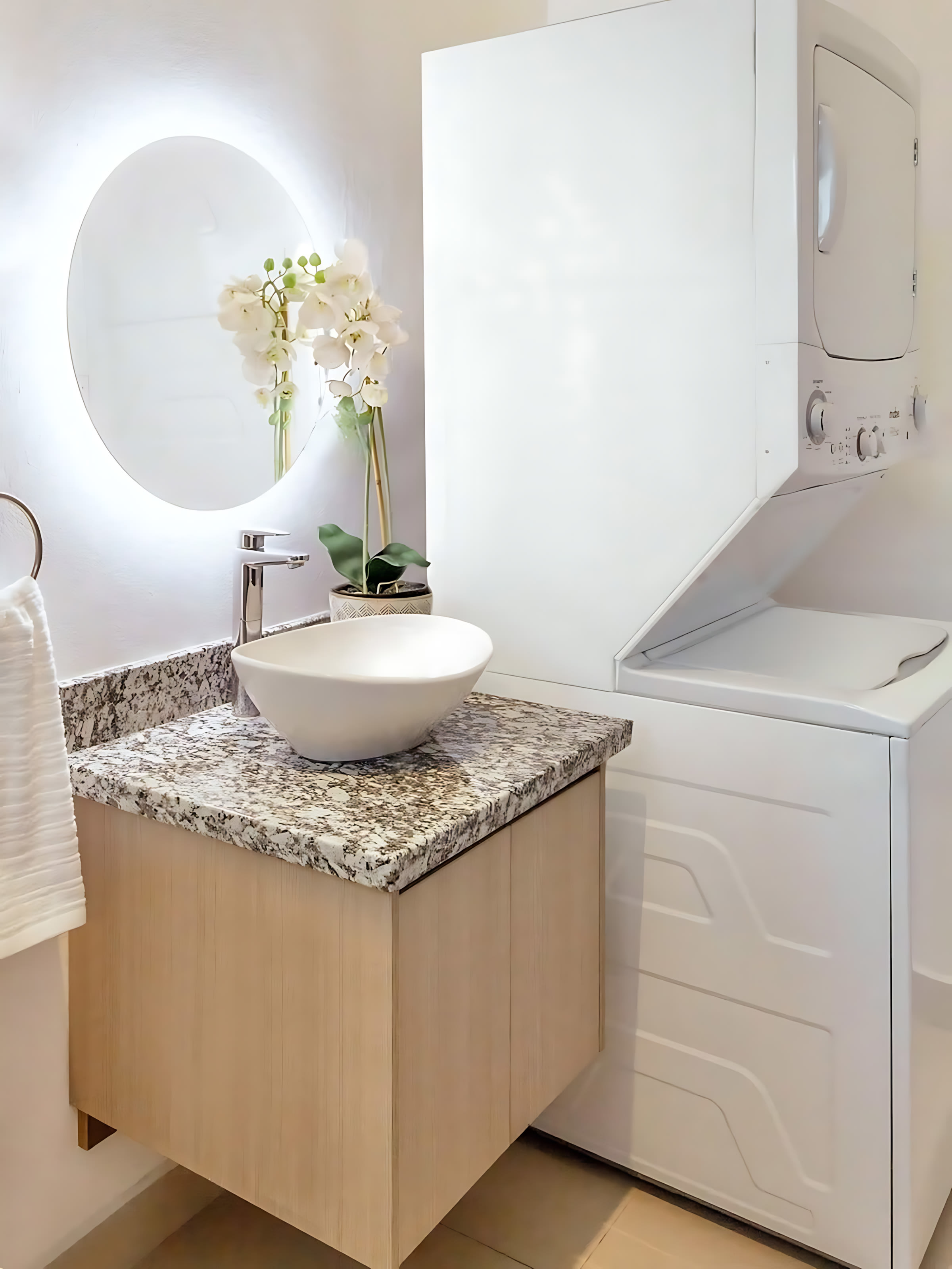 Modern bathroom featuring a white vessel sink on a speckled granite countertop, a light wood floating vanity, and a large backlit round mirror. A stacked white washer and dryer unit is visible to the right.
