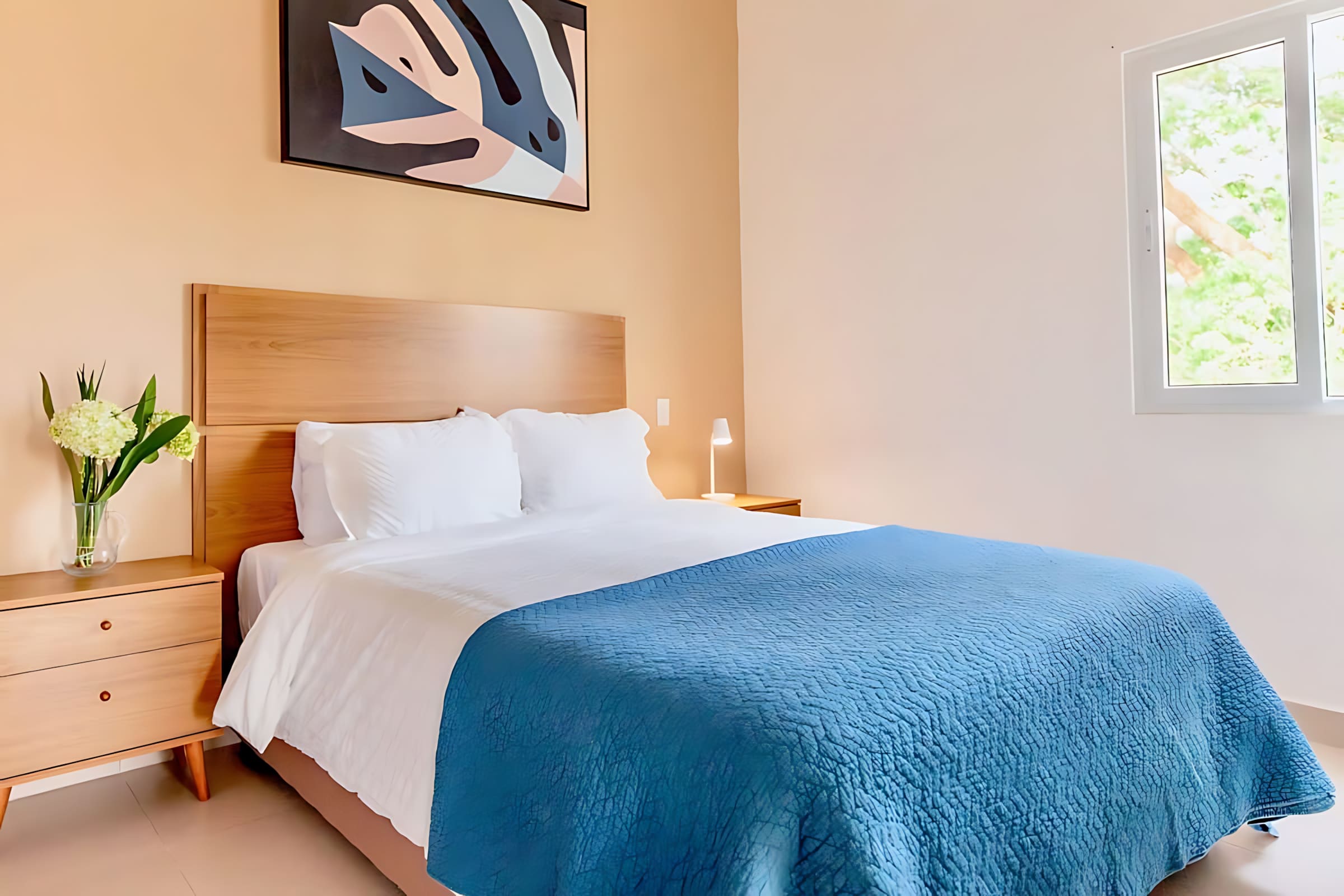 Bright bedroom with a queen-sized bed featuring a light wood headboard, white linens, and a blue quilted throw. An abstract artwork hangs above the bed, and a window provides natural light.