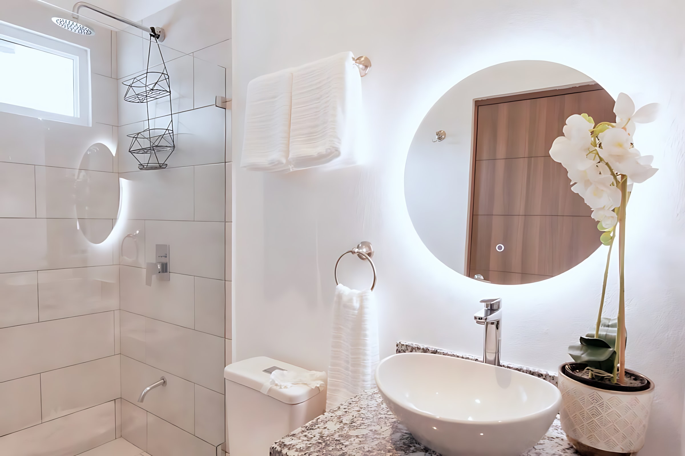 Modern bathroom with a white vessel sink on a granite-look vanity, illuminated by a large circular LED mirror. A walk-in shower with white subway tiles is visible to the left, and white towels are neatly folded on a rack.