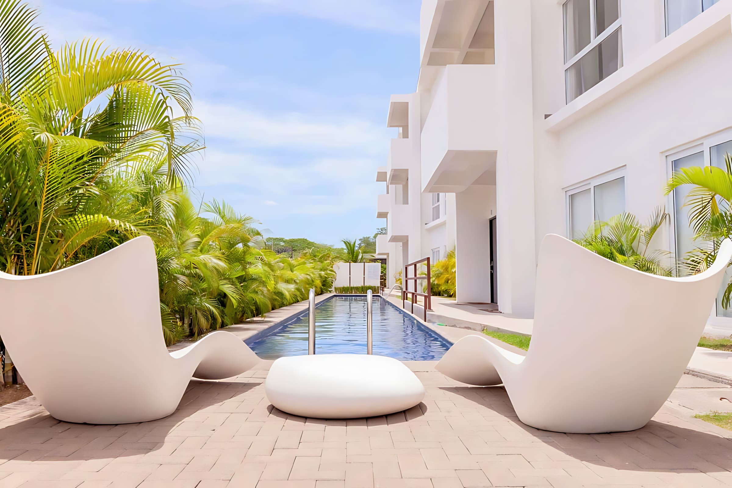Relaxing poolside area with two modern white lounge chairs and a matching low table on a paved patio. The long, narrow swimming pool is visible, flanked by vibrant green palm trees and the white facade of the building.