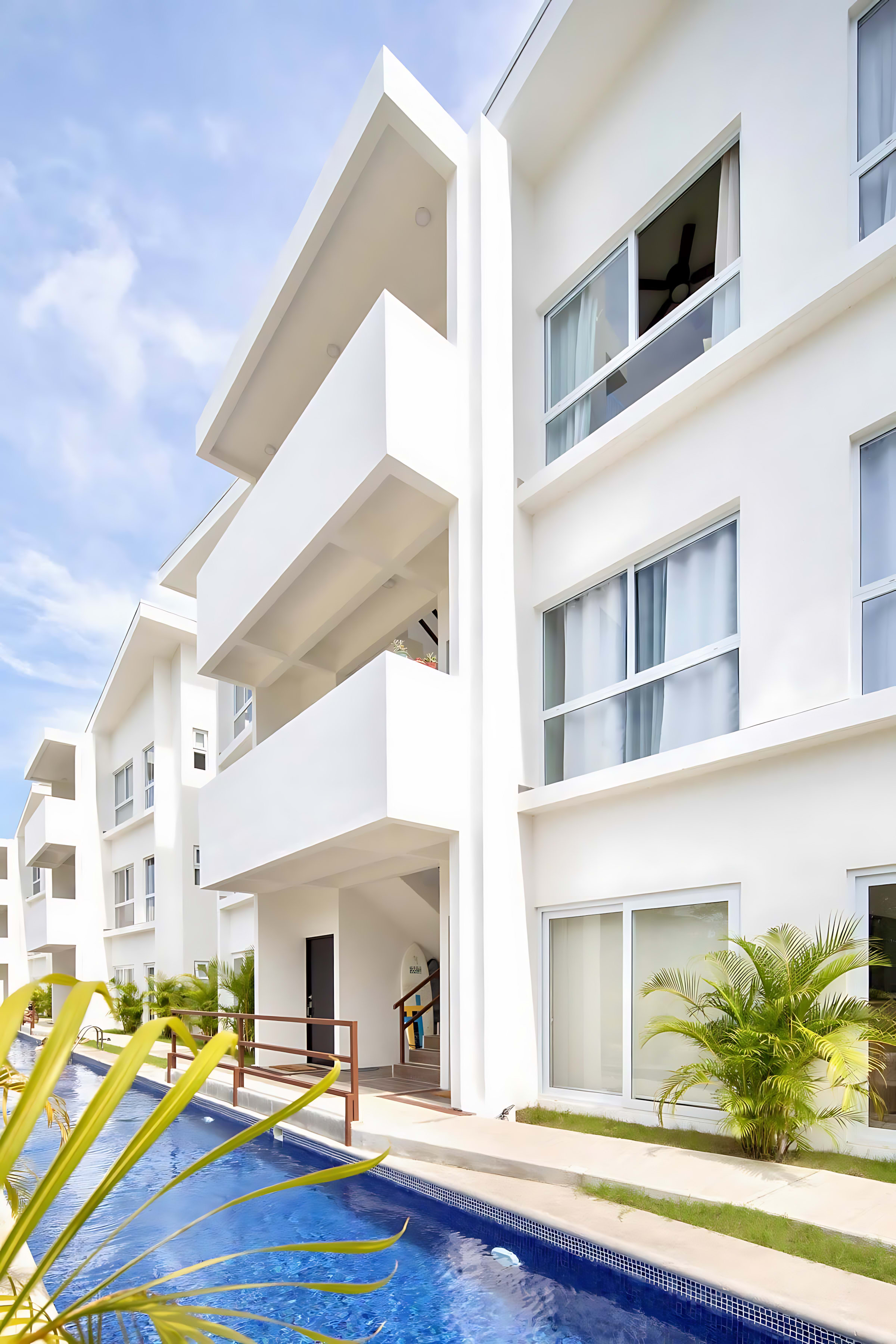 Modern white condominium buildings with balconies overlooking a long, narrow swimming pool with blue tiled bottom. Lush green and yellow palm trees line the pool area under a bright sky.