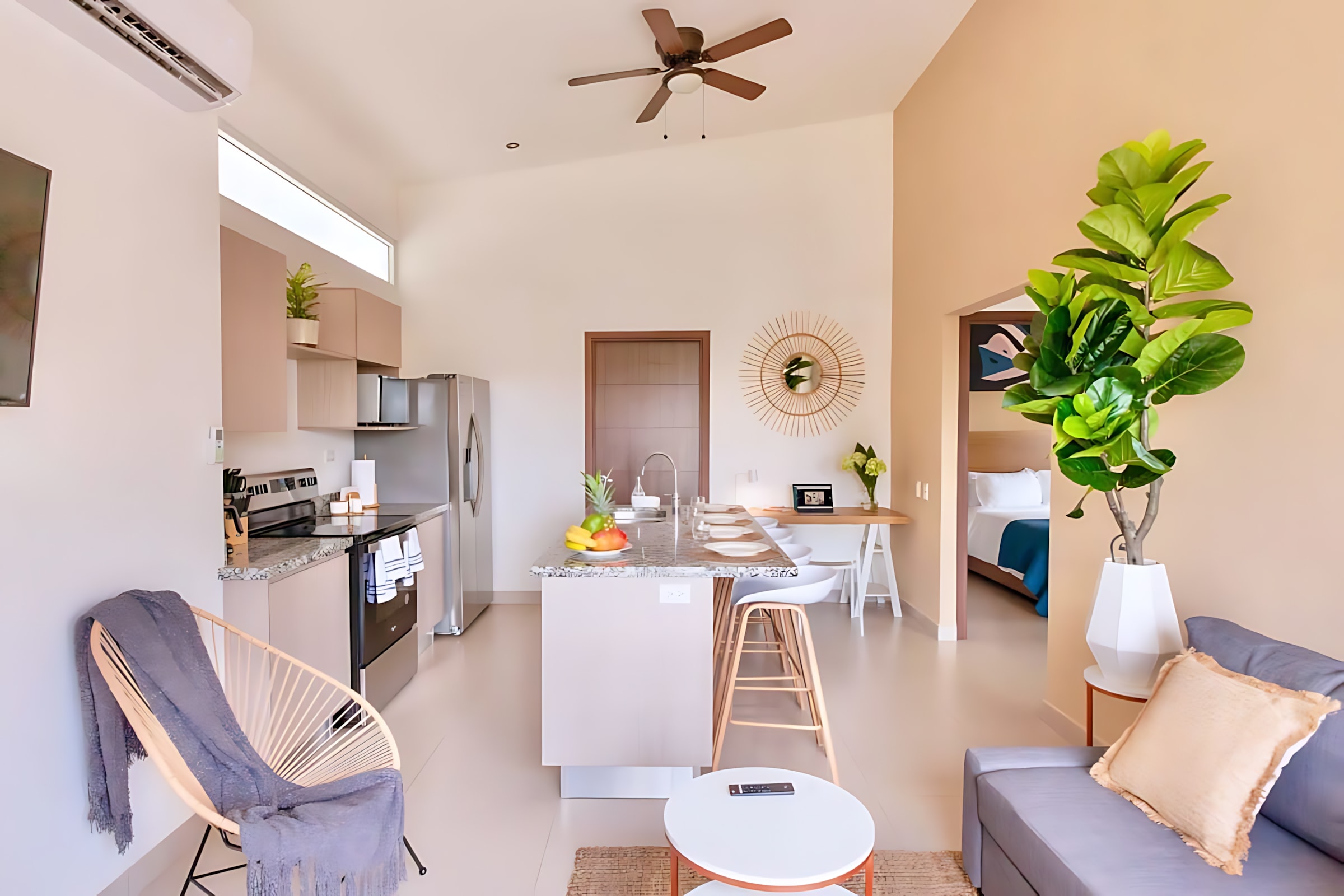 Spacious open-concept living room and kitchen with high ceilings. A grey sectional sofa faces a kitchen island with bar stools, and a large potted plant stands in the corner. A ceiling fan and air conditioning unit are visible.