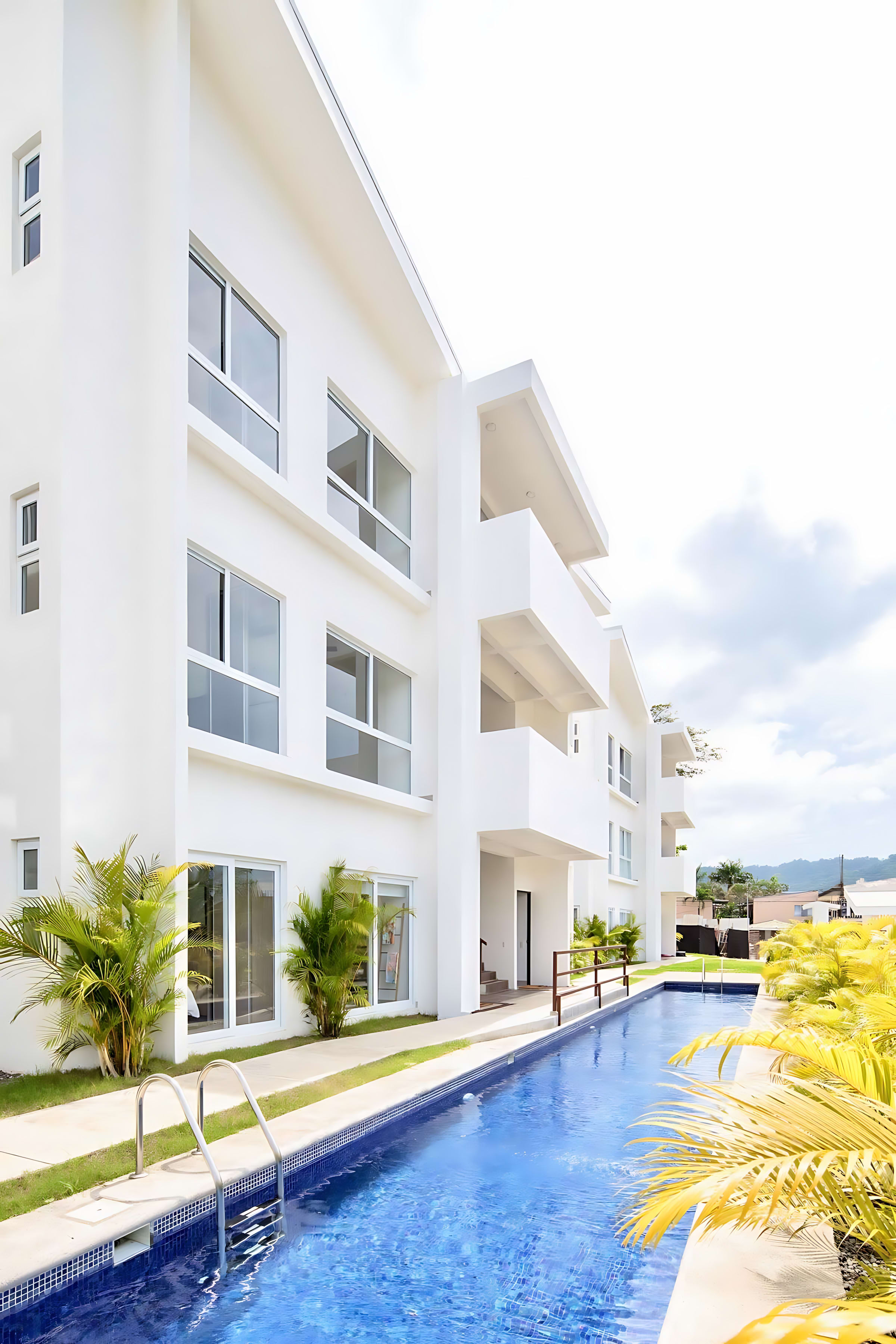 Expansive view of modern white condominium buildings with balconies, lining a long, narrow swimming pool. Green and yellow palm trees are visible along the pool's edge.
