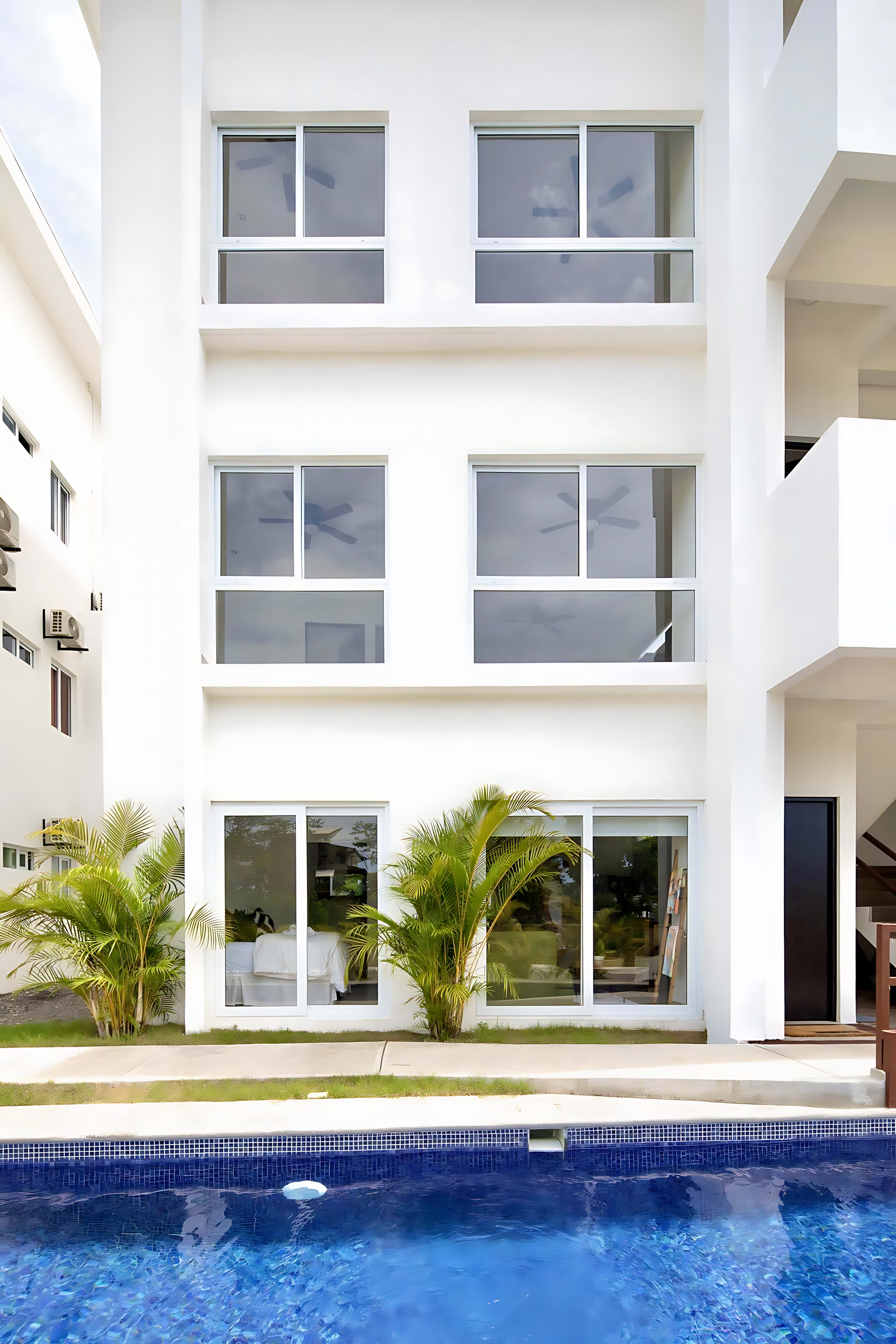 Front view of a modern white condominium building featuring large ground-floor windows and upper-level windows, situated directly beside a sparkling blue swimming pool.