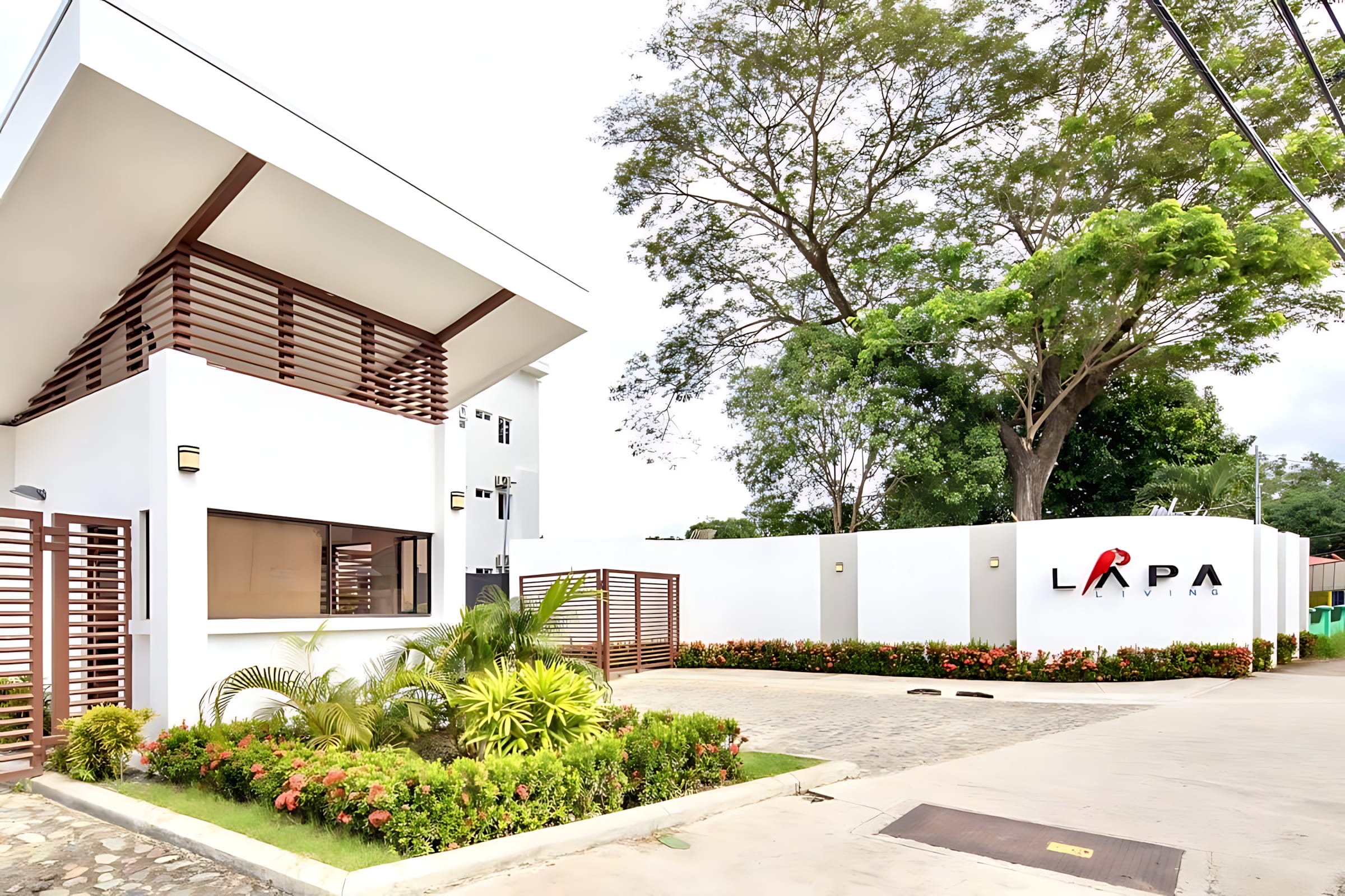Modern white building with brown slatted architectural details, surrounded by lush green landscaping and a white wall featuring the "LAPA LIVING" logo. A large mature tree stands tall in the background.
