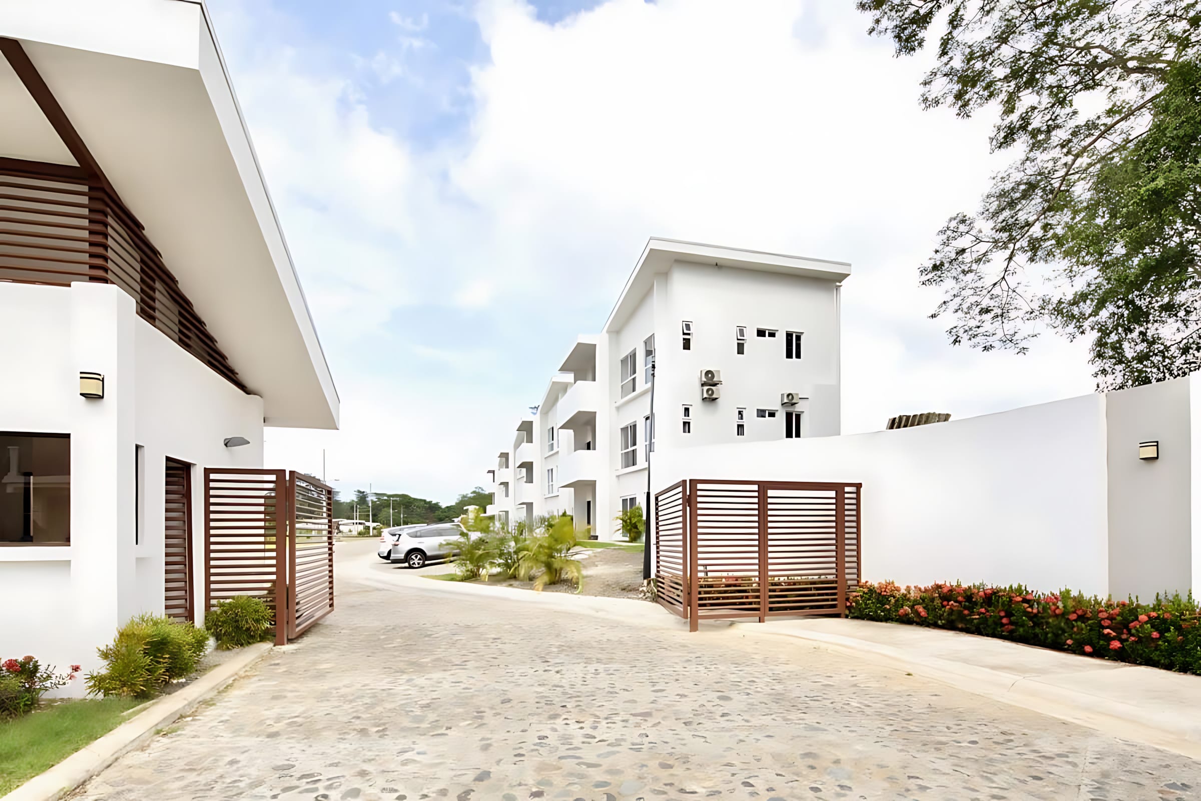 Paved entrance driveway leading into a modern residential complex with multiple white buildings featuring brown slatted gates and lush green landscaping. Parked cars are visible further down the drive.