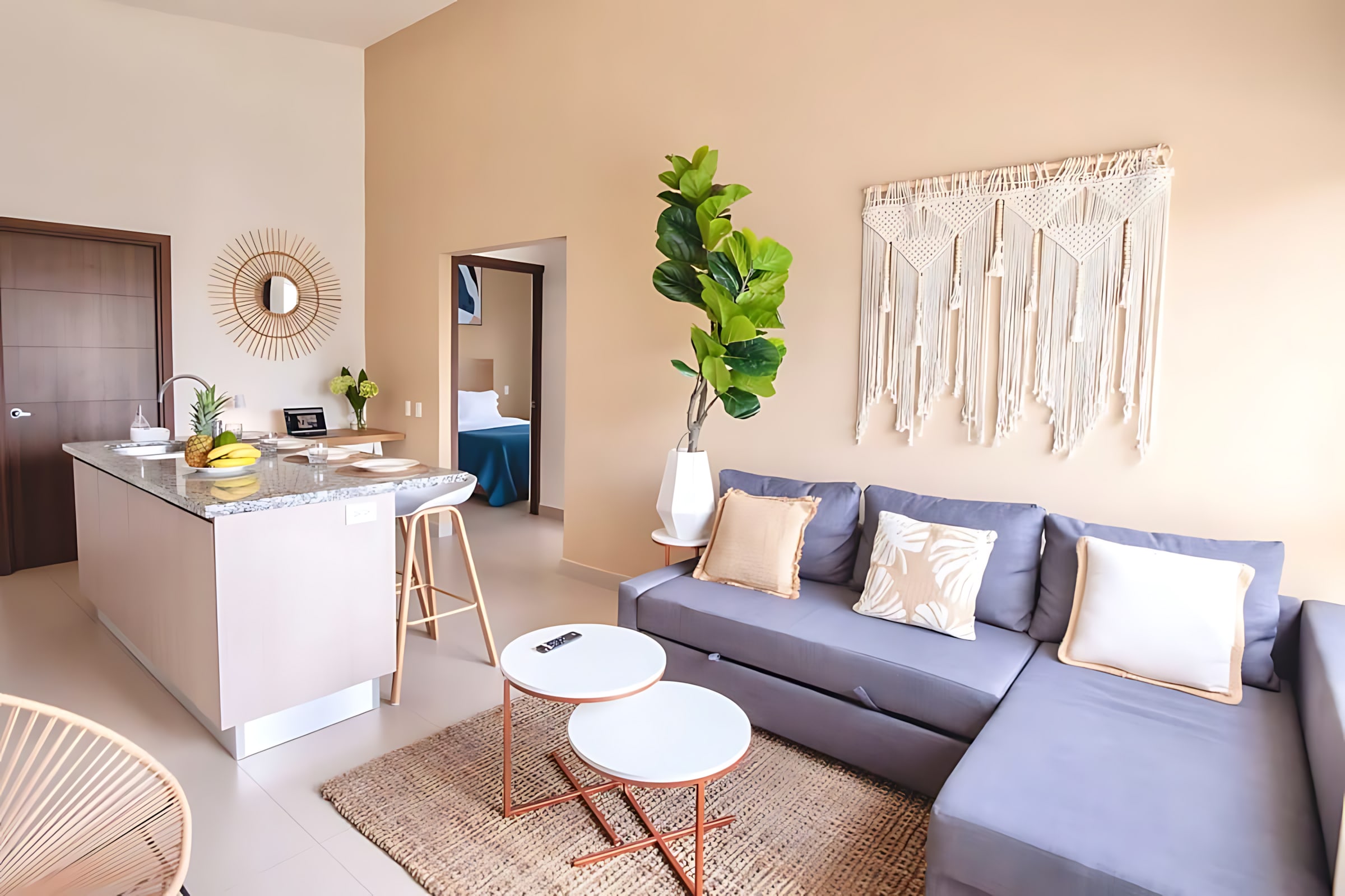View of the living room and kitchen from a different angle, highlighting the grey sectional sofa with decorative pillows and the kitchen island. A macrame wall hanging adorns the wall above the sofa, adding a bohemian touch.