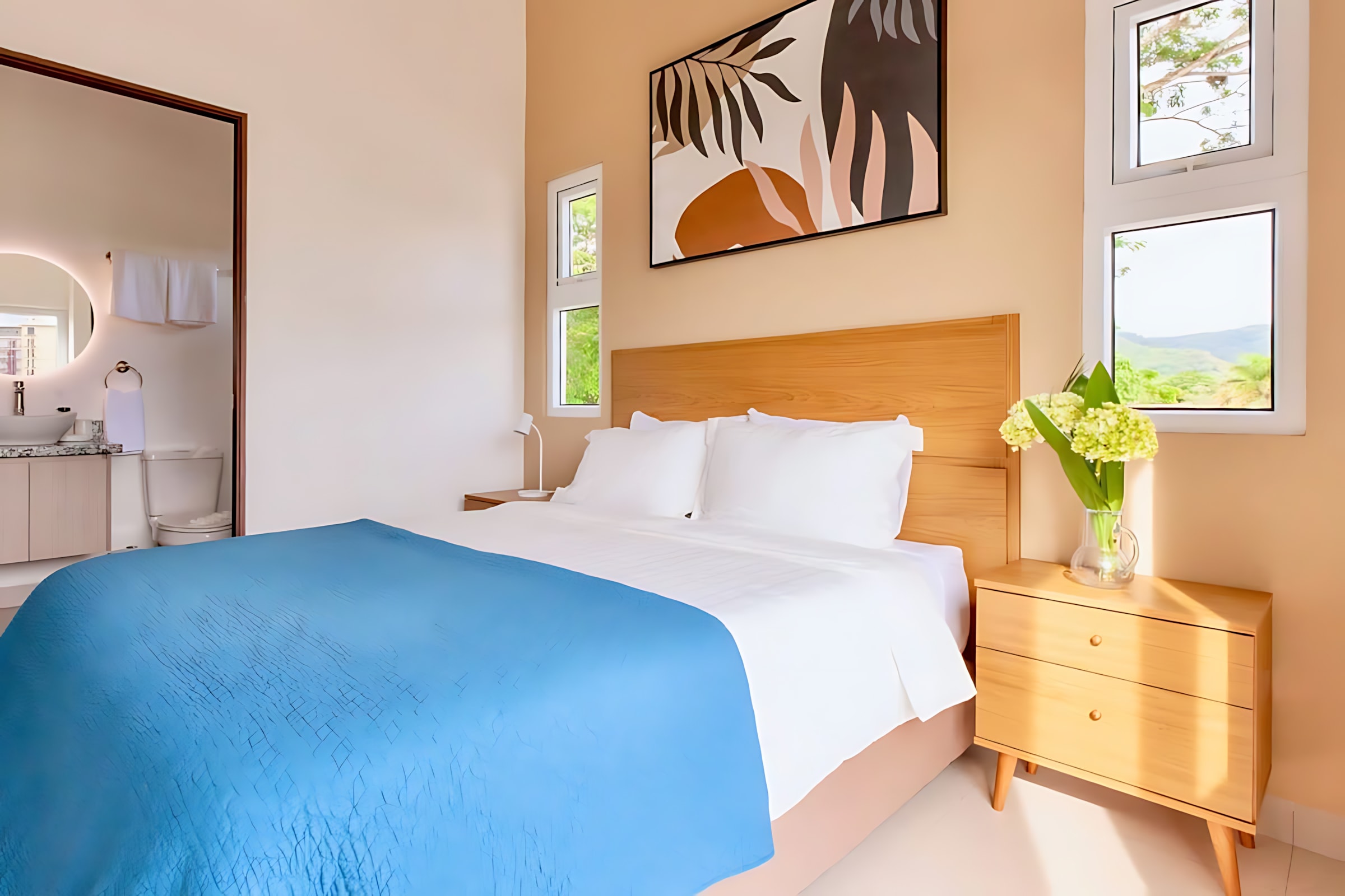 Bright bedroom with a wooden headboard bed, white bedding, and a blue quilted coverlet. A wooden nightstand with a vase of flowers is on the right, and a glimpse of the bathroom with a circular LED mirror is visible through the open doorway.
