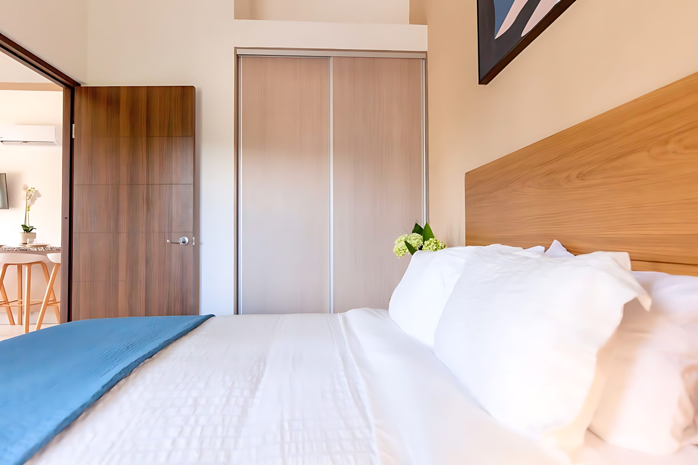 View from the bed in a modern bedroom, showing light wood sliding closet doors and a dark wood door leading to another room. White pillows and bedding are visible in the foreground.