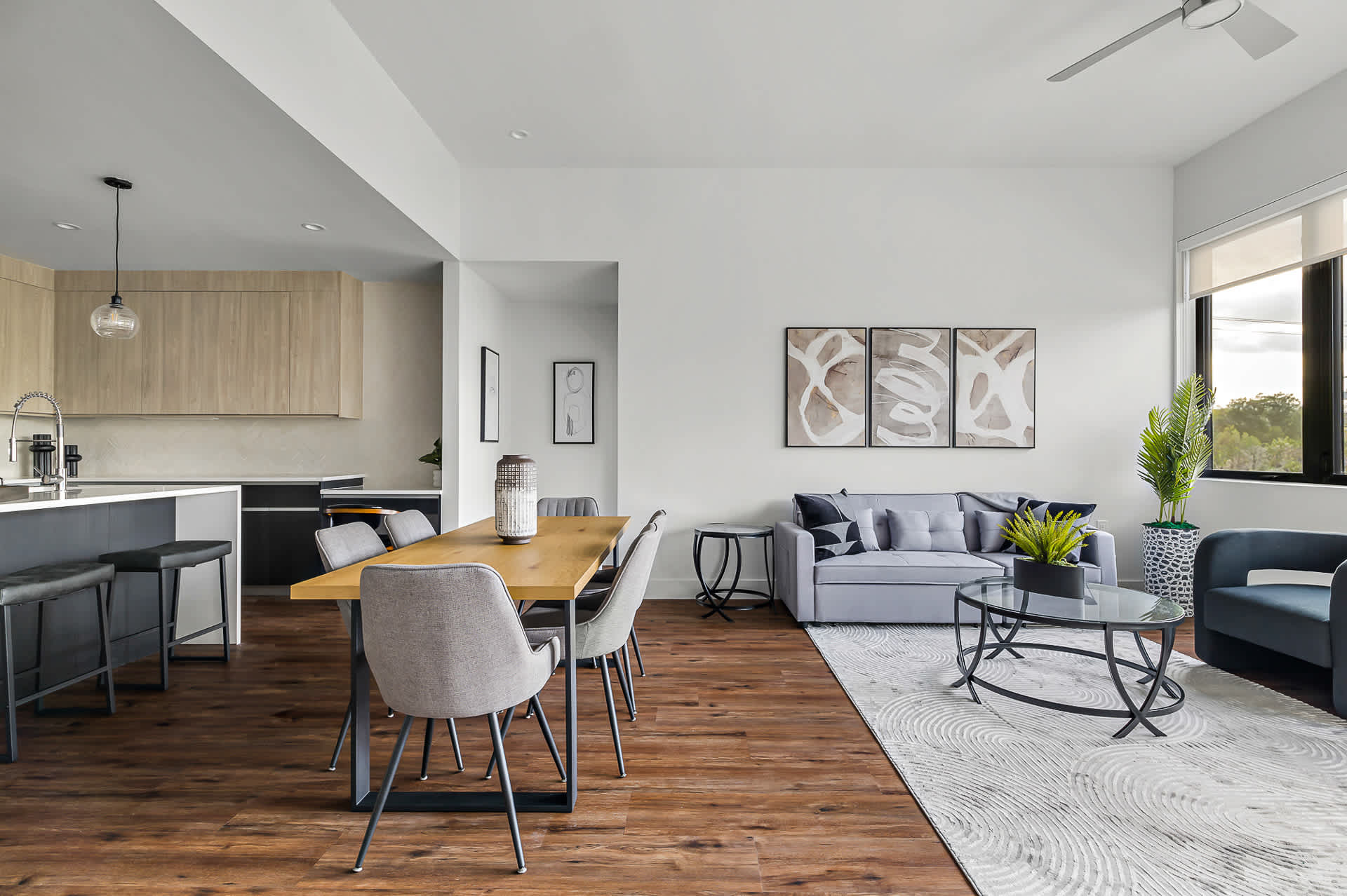 Expansive open-concept living and dining area featuring rich hardwood floors, a light gray sofa, and a wooden dining table with gray upholstered chairs. Large windows offer views of green trees, and modern abstract art adorns the wall.