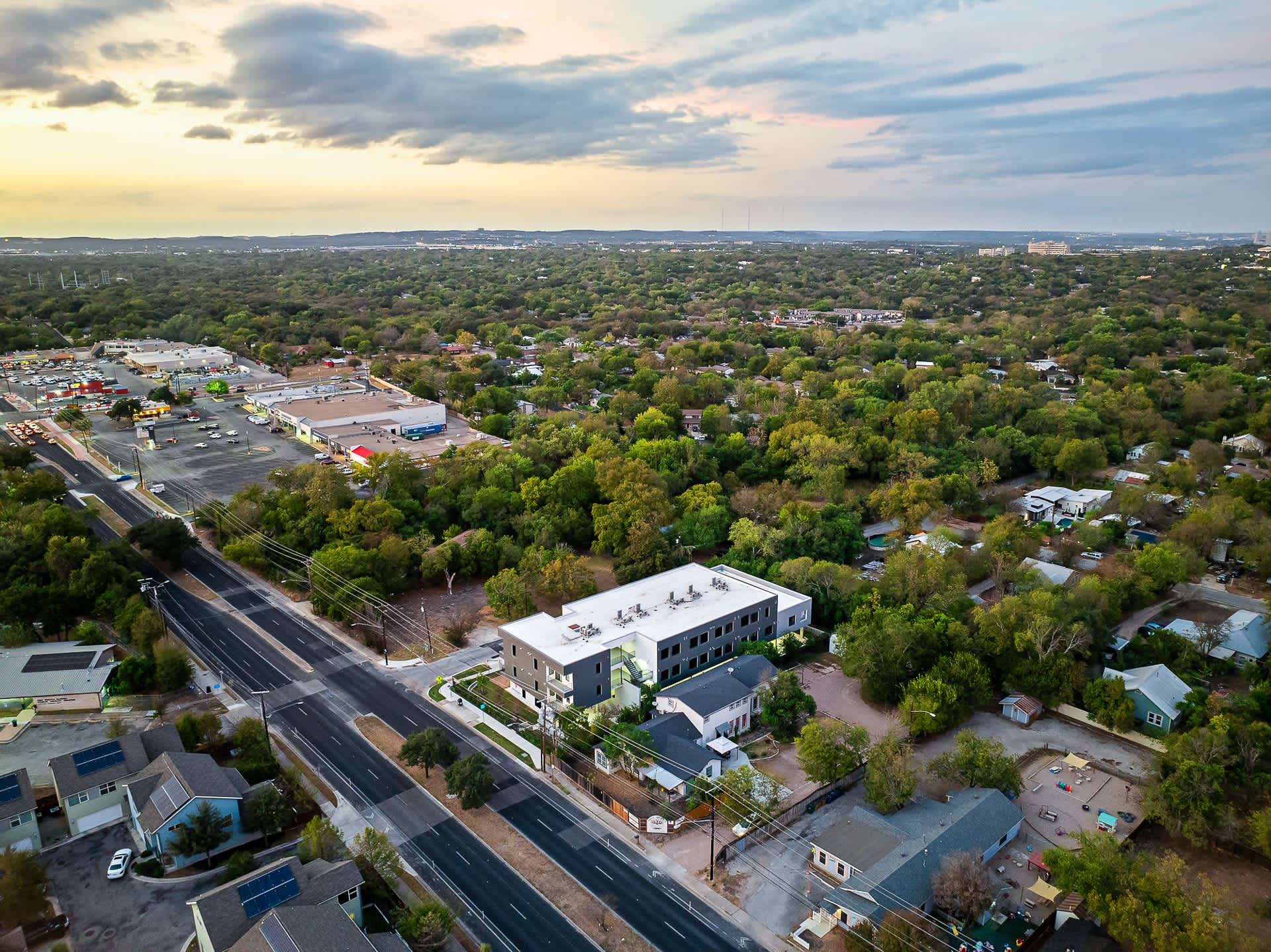 A broad aerial perspective shows the apartment building alongside a main road, surrounded by a mix of residential homes and commercial establishments, all set within a verdant landscape under a cloudy sky.