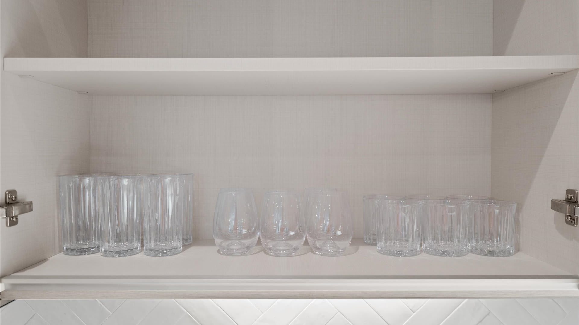 An open kitchen cabinet showcasing a collection of clear glass drinkware, including tall ribbed glasses, stemless wine glasses, and shorter tumblers, neatly organized on a shelf.