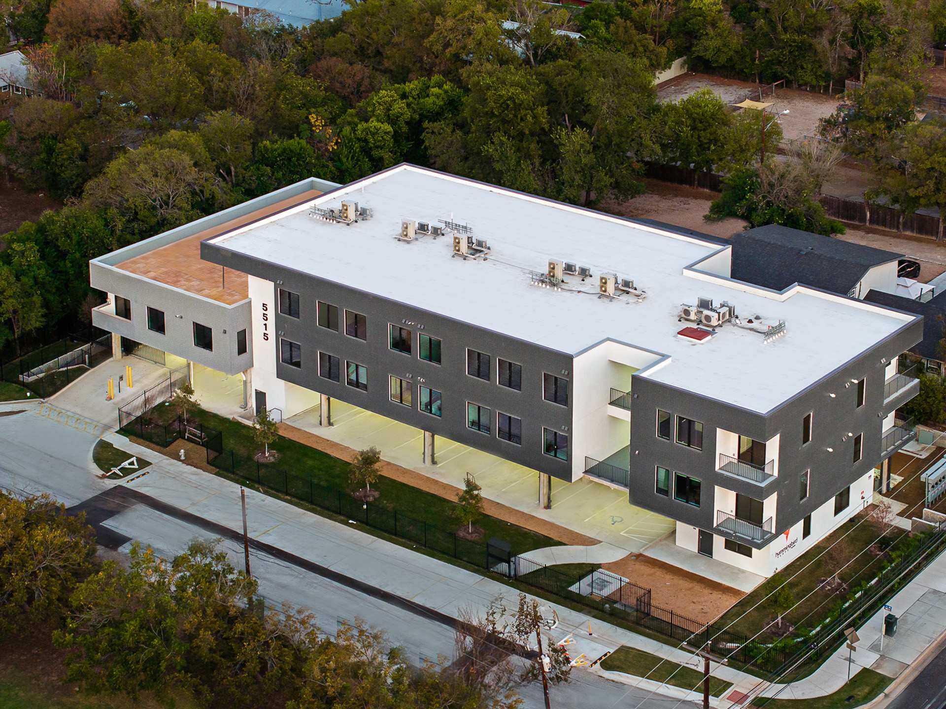 An aerial view showcases the modern, multi-level apartment building with a flat white roof and dark gray facade, surrounded by paved parking areas, green lawns, and mature trees under a bright sky.