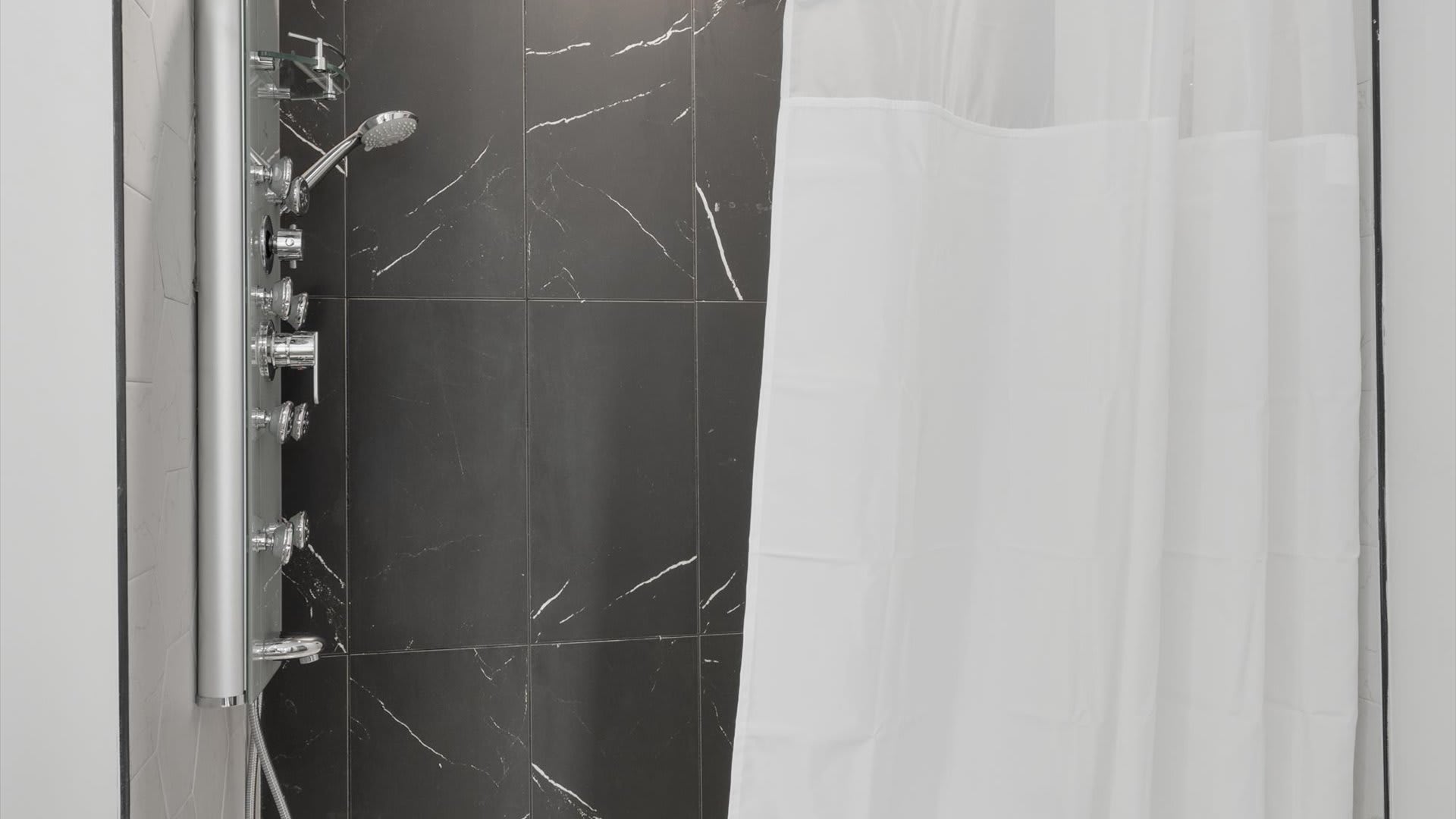 Close-up of the walk-in shower in bathroom-1, showcasing black marble-look tiles and a modern multi-function shower panel with various spray options. A white shower curtain is partially drawn.