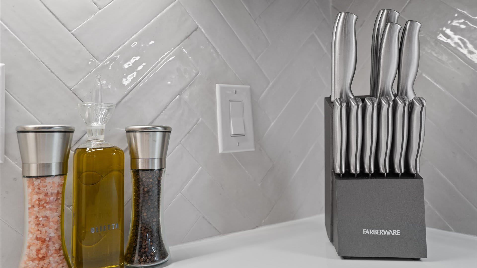 A close-up of a kitchen countertop featuring a gray knife block filled with silver-handled knives, alongside a clear glass cruet of olive oil, a salt grinder, and a pepper grinder. A white herringbone tile backsplash and a light switch are visible behind.