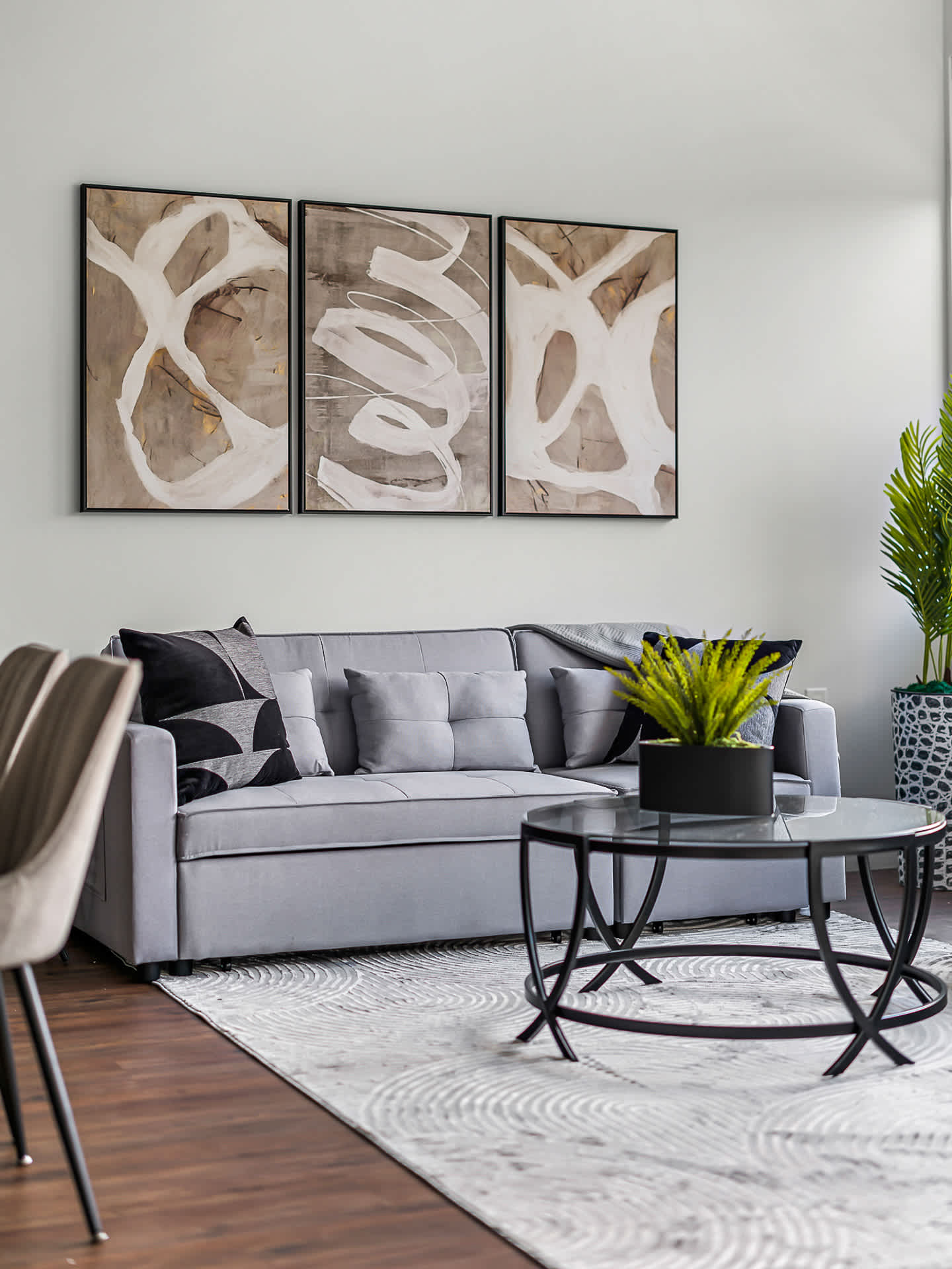 Close-up of a modern living room featuring a light gray sectional sofa with decorative pillows, a round glass coffee table, and a light-colored patterned area rug. Three framed abstract art pieces hang above the sofa, adding a contemporary touch.
