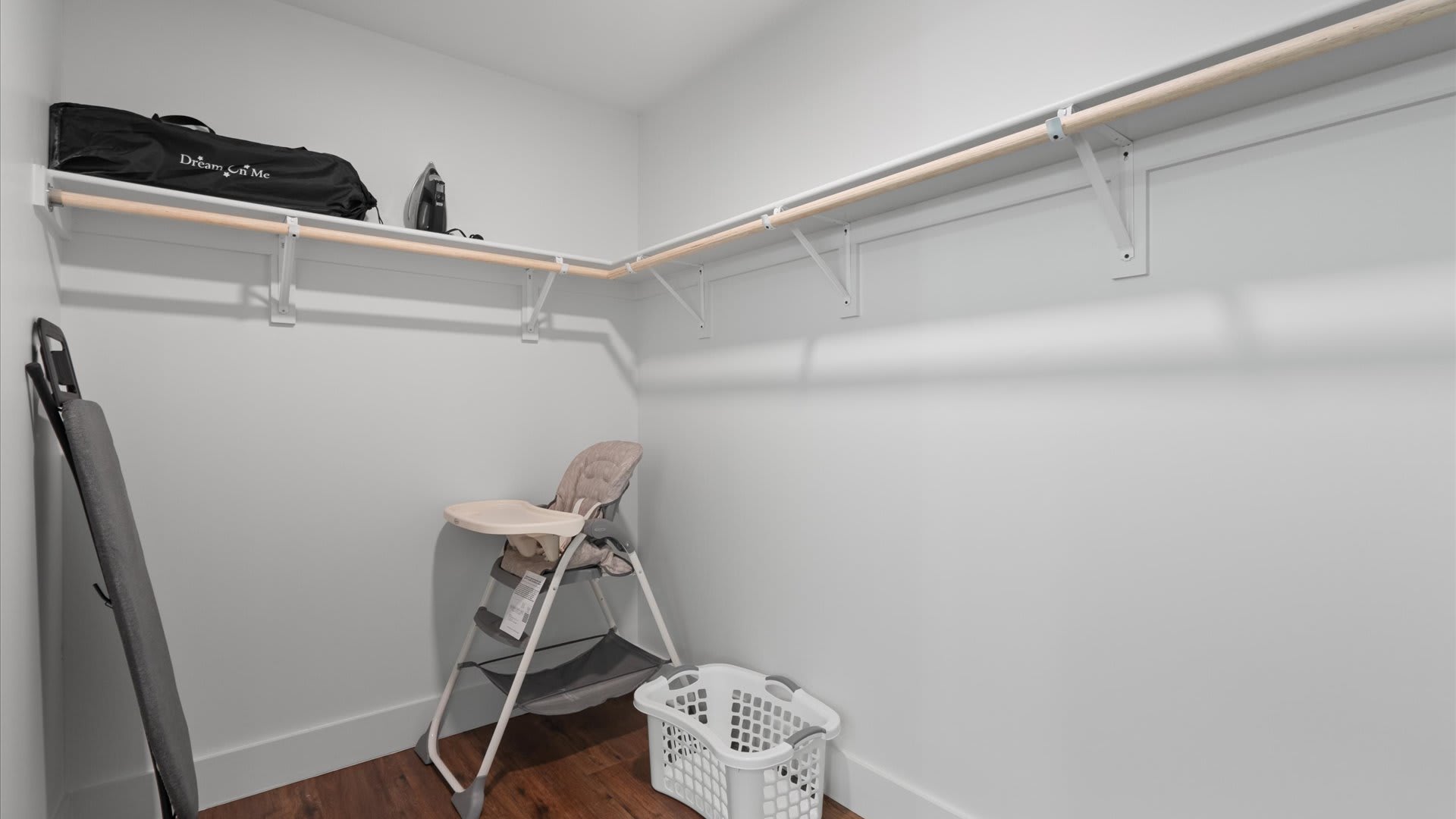 A spacious walk-in closet with light gray walls and wood-look flooring features white wire shelving and hanging rods, holding a portable high chair, a laundry basket, and a "Dream On Me" travel crib bag.