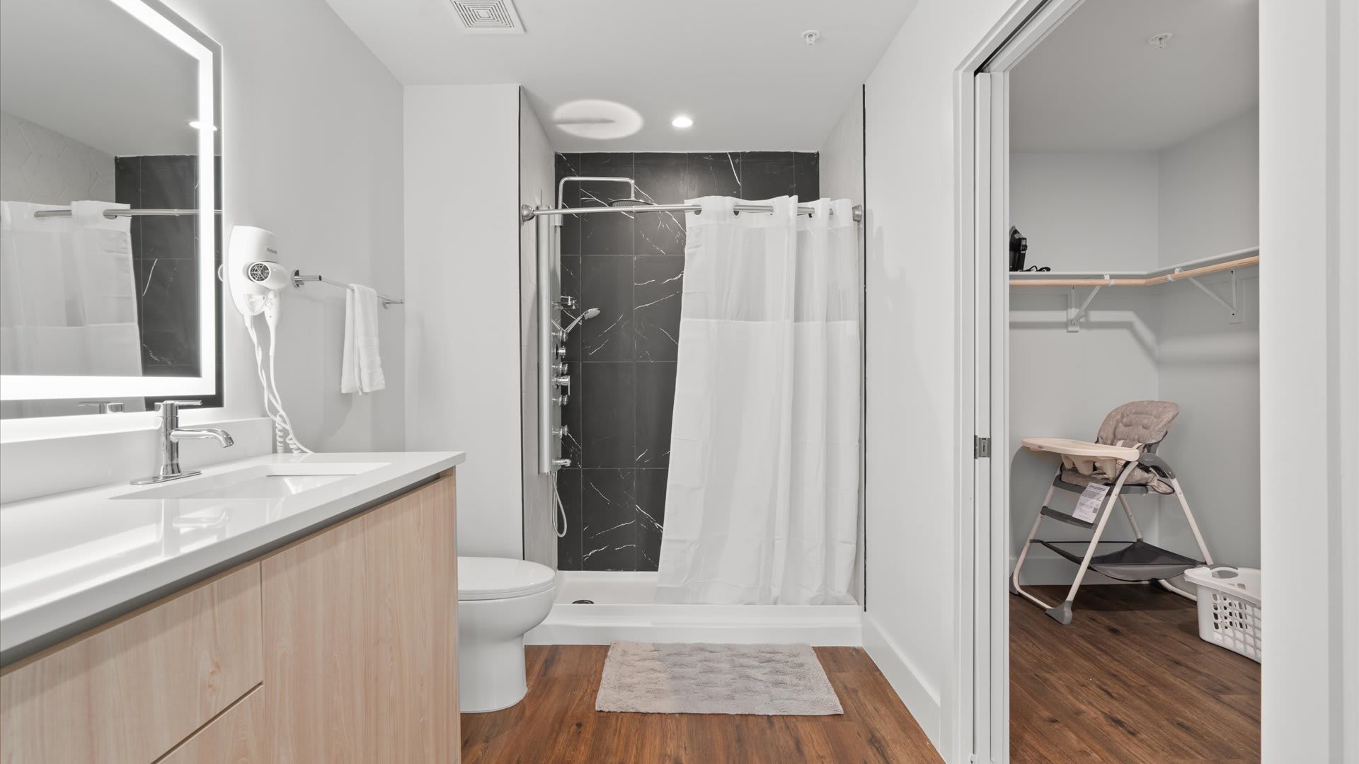Wide view of bathroom-1, showing the double vanity, toilet, and walk-in shower with black marble-look tiles. An open doorway reveals a closet with shelving and a high chair, indicating family-friendly amenities.