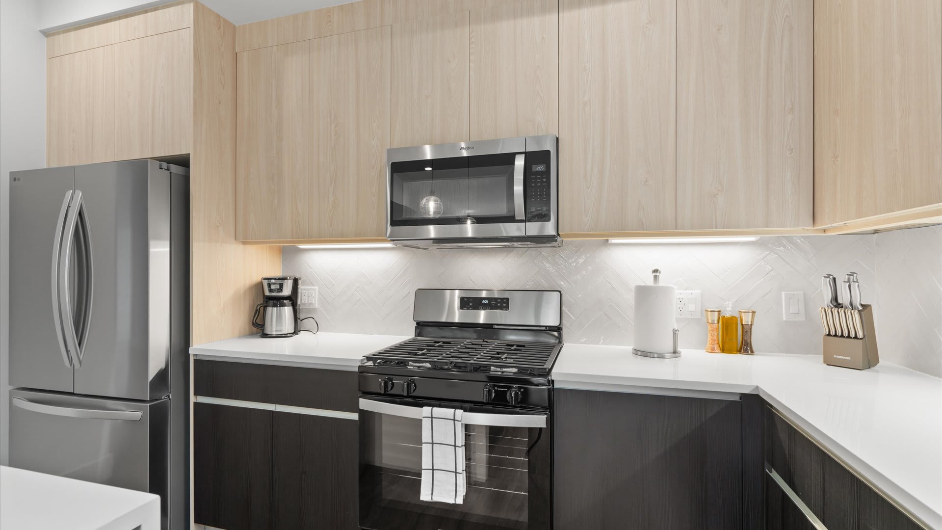 A modern kitchen featuring light wood upper cabinets and dark wood lower cabinets, white quartz countertops, a stainless steel gas range with an oven, a matching microwave above, and a large stainless steel refrigerator. A white herringbone tile backsplash adds texture, and under-cabinet lighting illuminates the workspace.