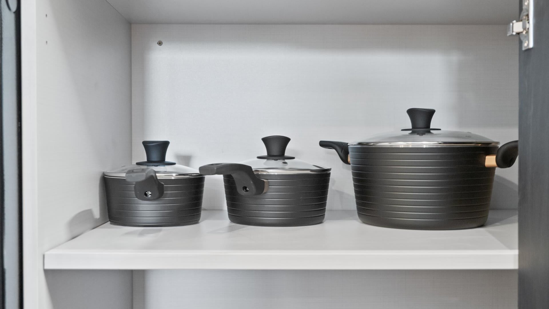 A kitchen cabinet shelf neatly arranged with three dark gray non-stick pots of varying sizes, each with a glass lid and black handles, ready for cooking.