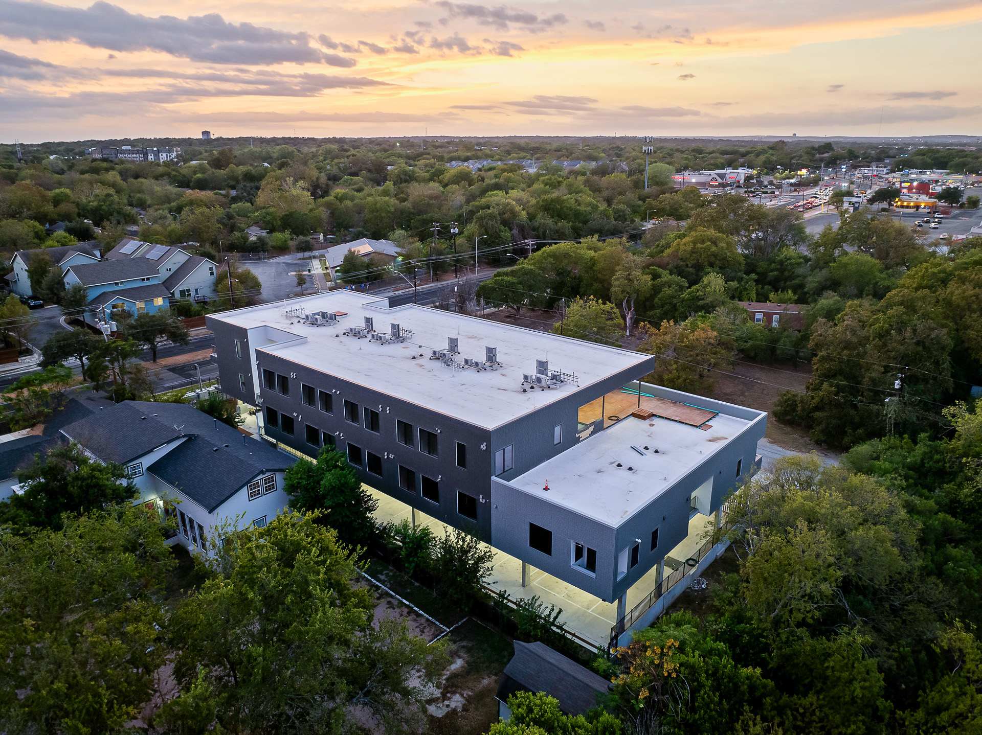 An aerial shot at dusk reveals the modern apartment building with its flat roof and dark facade, nestled among lush green trees, with a soft orange and purple sunset sky in the background.