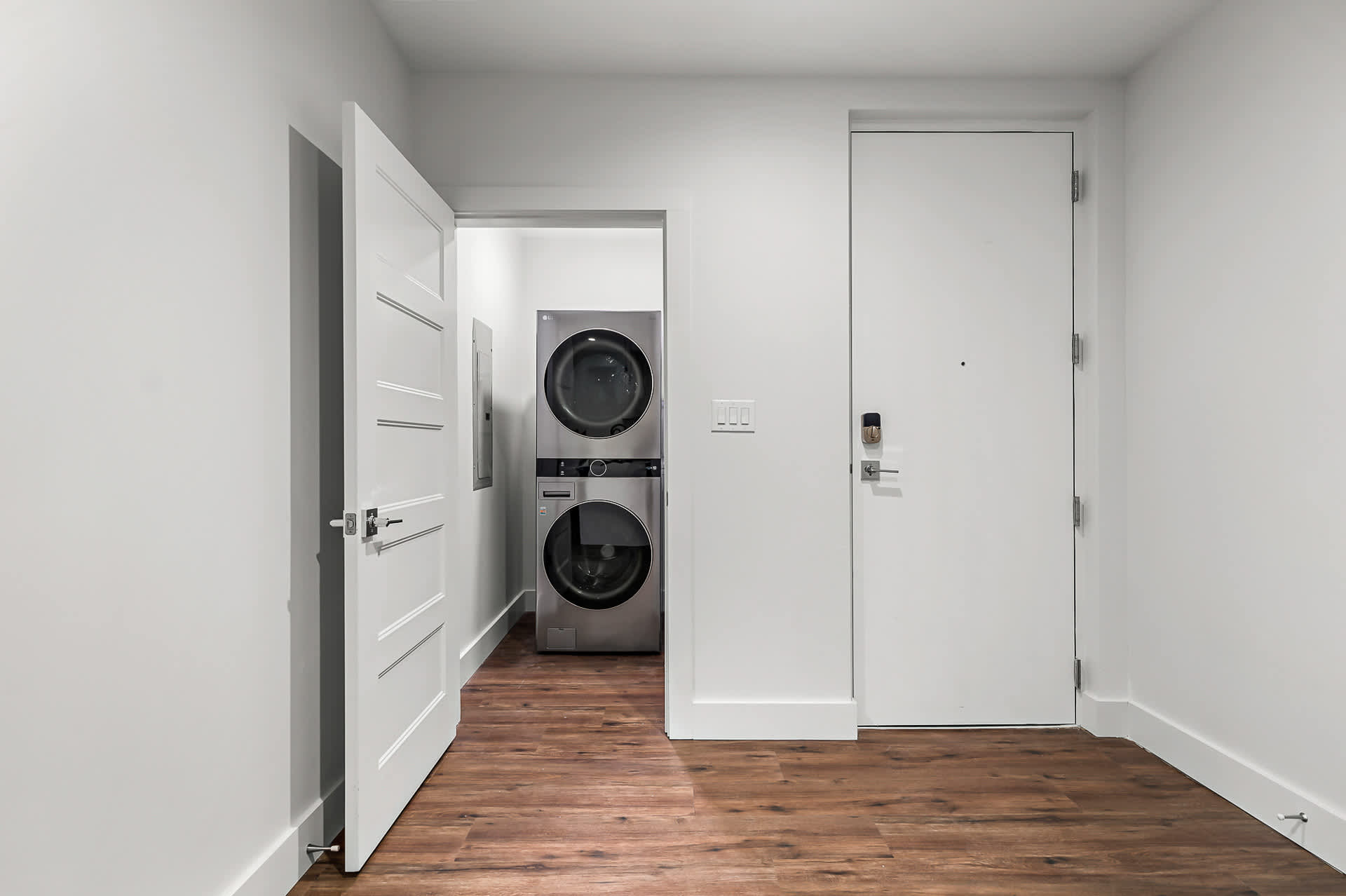 A bright hallway with light wood-look flooring leads to a laundry closet featuring a stacked silver washer and dryer, visible through an open white door. Another white door with a smart lock is on the right.