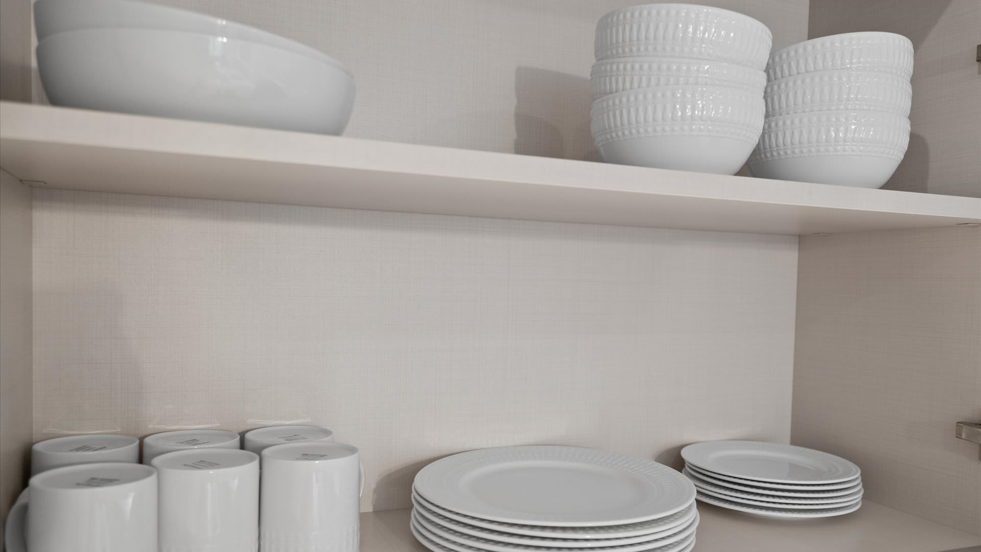 A well-stocked kitchen cabinet shelf featuring white ceramic dinnerware, including stacks of plates, bowls with textured exteriors, and mugs, all neatly arranged.