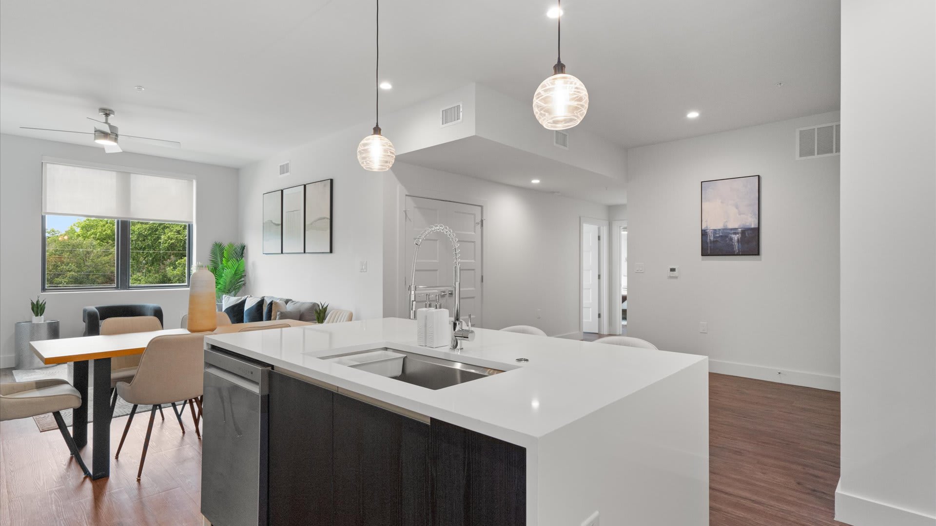 An open-concept living space featuring a modern kitchen island with a sink and dishwasher, leading into a dining area with a light wood table and chairs, and a living room with a sofa and large window. The space has light wood flooring, white walls, and two clear glass pendant lights over the island.