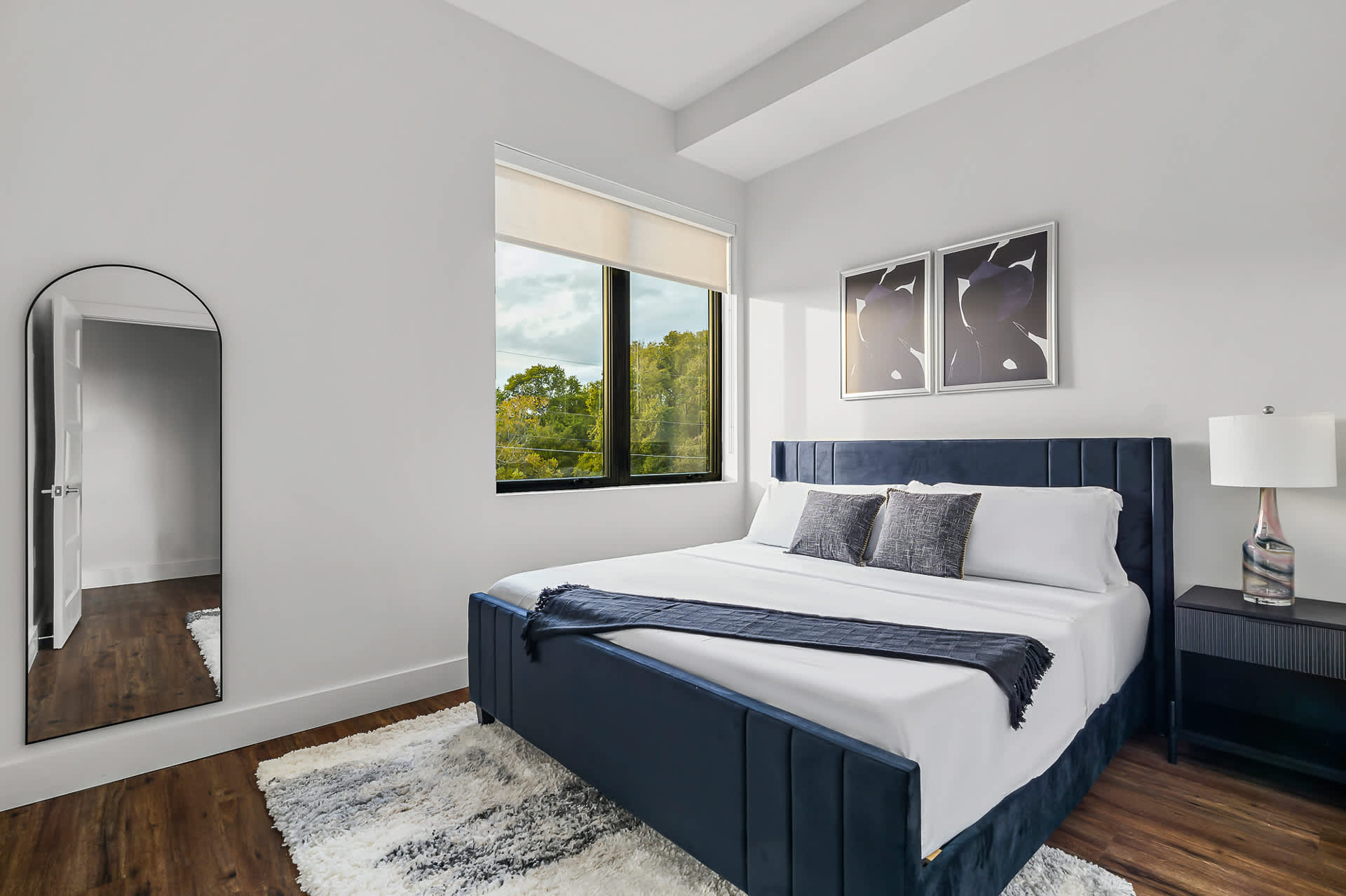 Wider view of the second bedroom, showcasing the navy blue upholstered bed, a fluffy white area rug, and a tall arched full-length mirror on the left. A large window with a roller shade provides ample natural light and a view of trees.