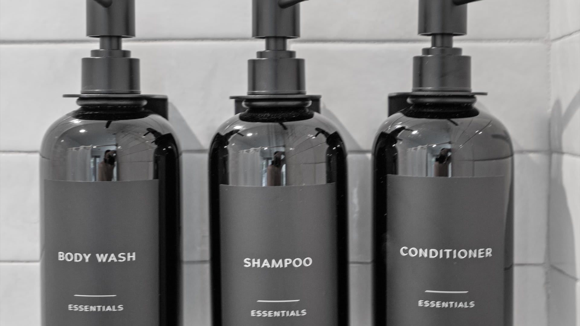 Close-up of three black pump bottles labeled "BODY WASH", "SHAMPOO", and "CONDITIONER" mounted on a white tiled shower wall, indicating provided toiletries.