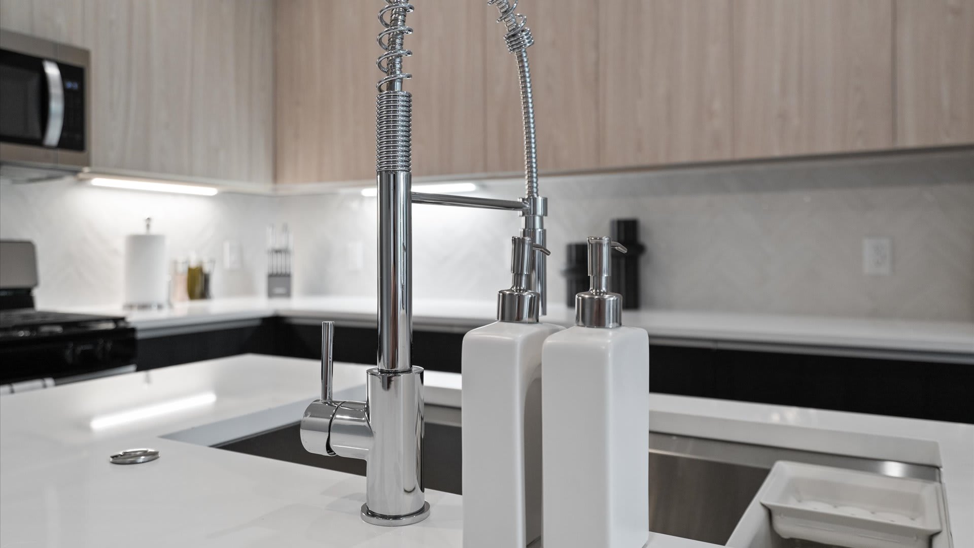 A close-up of a modern kitchen island with a stainless steel sink and a chrome pull-down faucet, flanked by two white rectangular soap dispensers. The white quartz countertop and light herringbone tile backsplash are visible in the background.