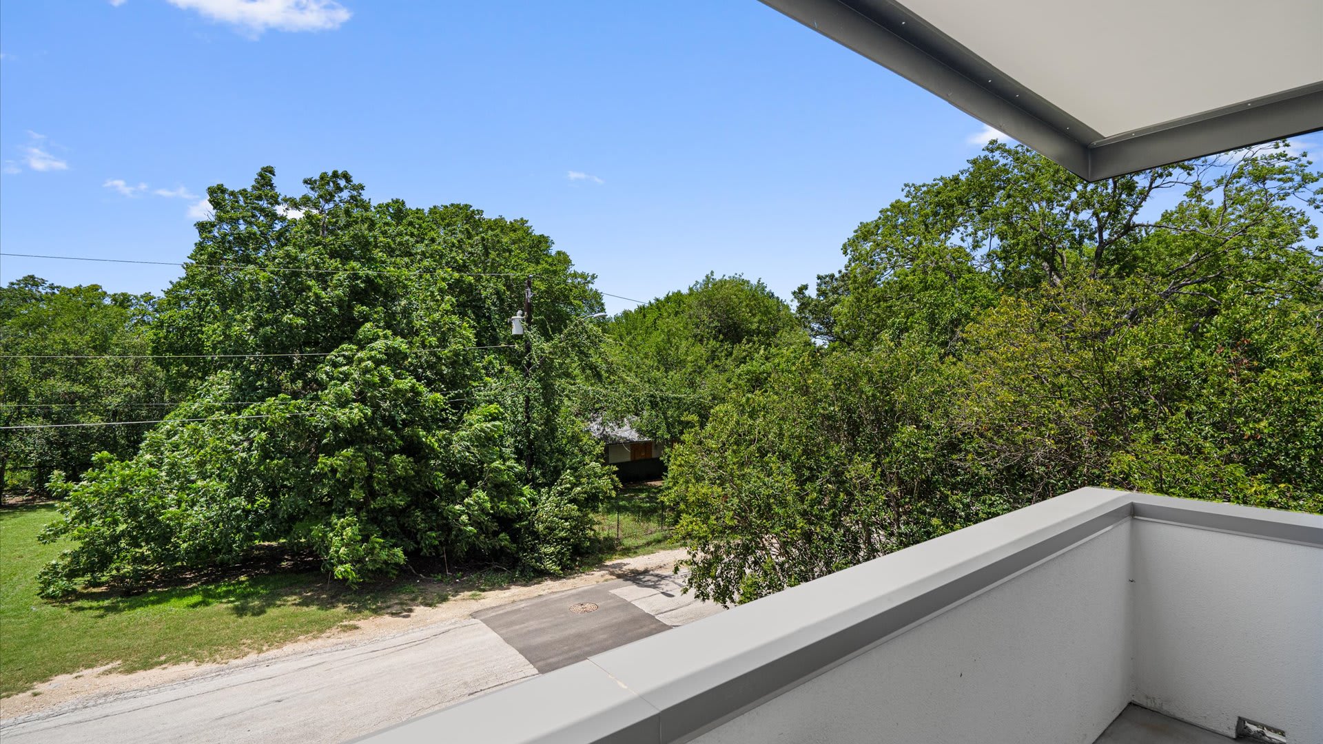 A scenic view from a balcony shows a canopy of dense green trees under a bright blue sky, with a narrow road visible below, suggesting a peaceful, natural setting.