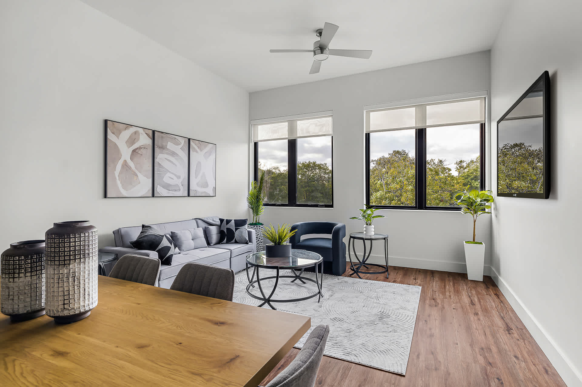 Bright and airy open-concept living and dining space with dark hardwood floors, a gray sofa, and a wooden dining table. Large windows provide ample natural light and views of surrounding trees, while a wall-mounted TV and ceiling fan complete the modern aesthetic.