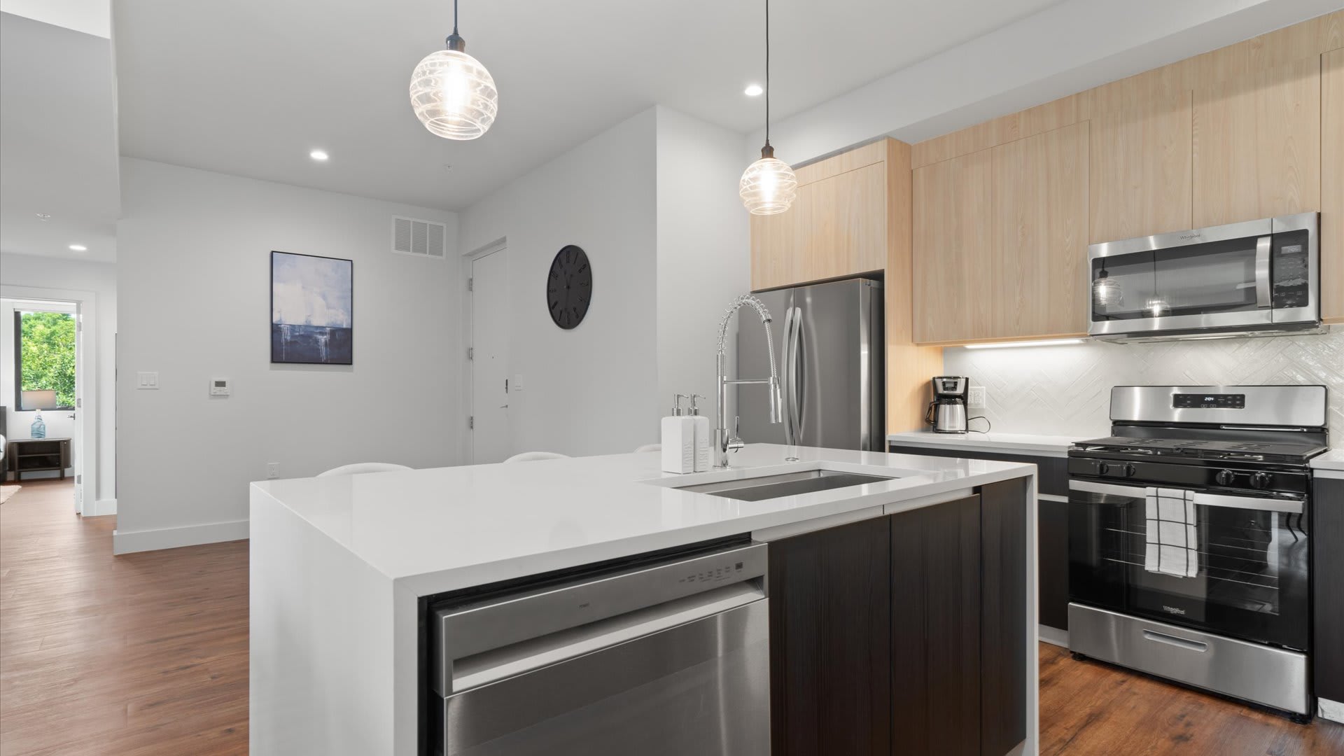 A wide shot of a contemporary kitchen with a large white quartz island featuring a stainless steel sink and dishwasher, dark wood cabinetry, and two clear glass pendant lights overhead. The kitchen connects to an open living area with light wood flooring and white walls.