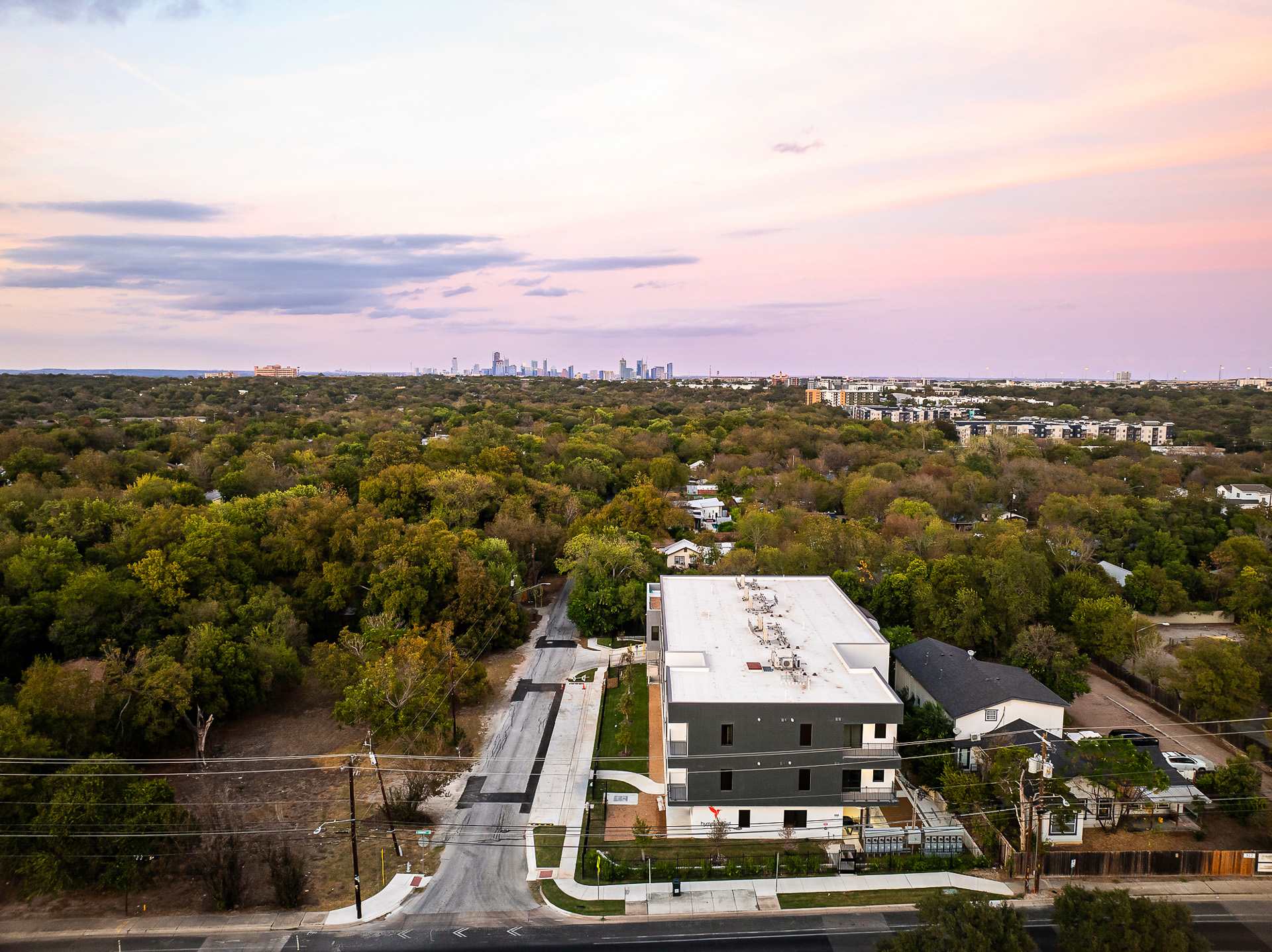 A wide aerial view captures the apartment building surrounded by a dense green tree canopy, with a distant city skyline visible under a soft pink and purple sky, highlighting its suburban setting.