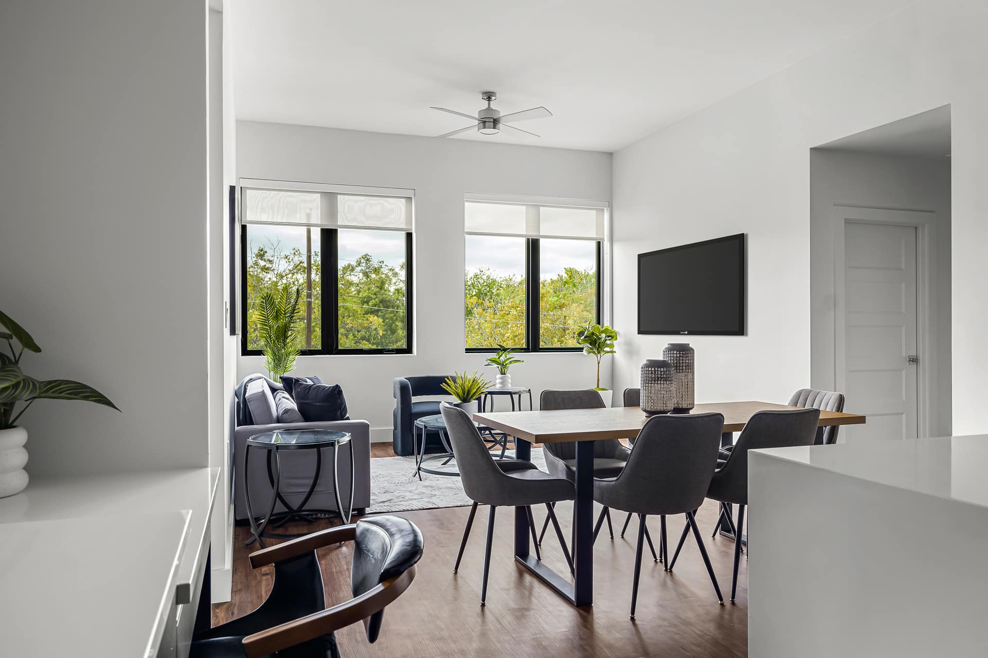 View of the open-concept living and dining area from the kitchen, showcasing the wooden dining table with gray chairs and the comfortable gray sofa. The space is well-lit by natural light from large windows, and a wall-mounted TV is visible.
