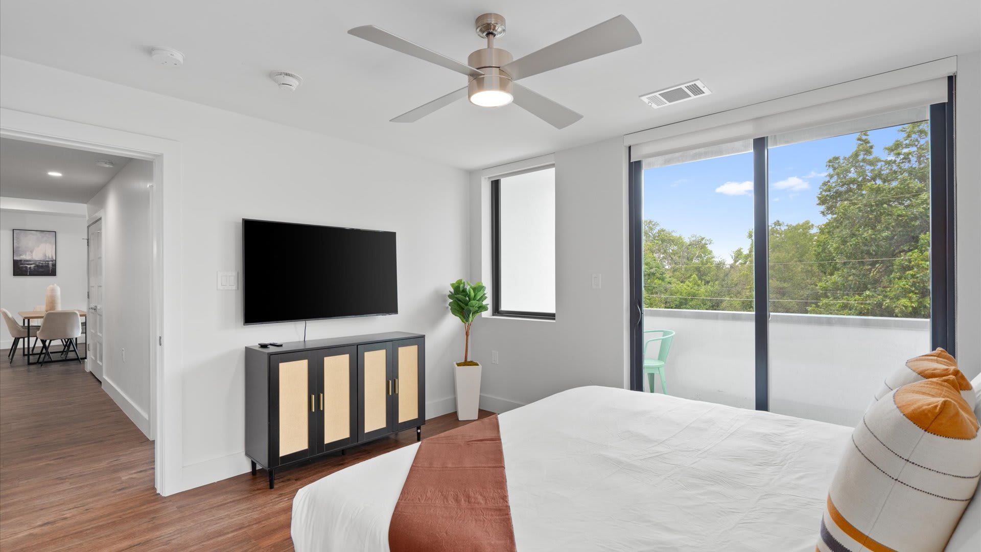 Modern bedroom with a large flat-screen TV mounted above a black and natural wood cabinet, a potted plant, and a ceiling fan. A large window and glass door lead to a balcony with a light green chair, overlooking lush trees.