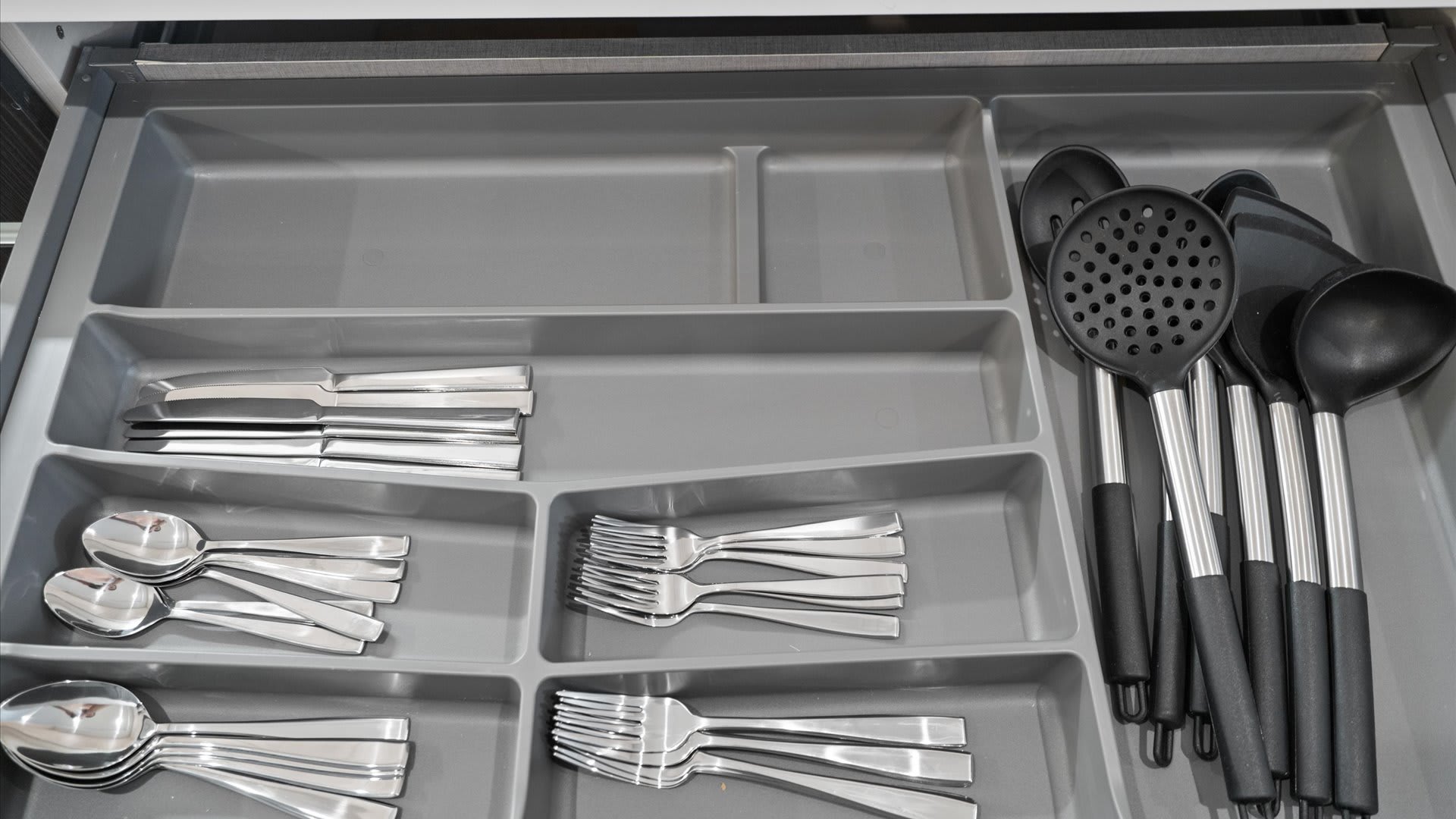 A close-up view inside a kitchen drawer showing a gray plastic organizer filled with various silver cutlery, including forks, spoons, and knives, alongside black-handled serving utensils like ladles and a slotted spoon.