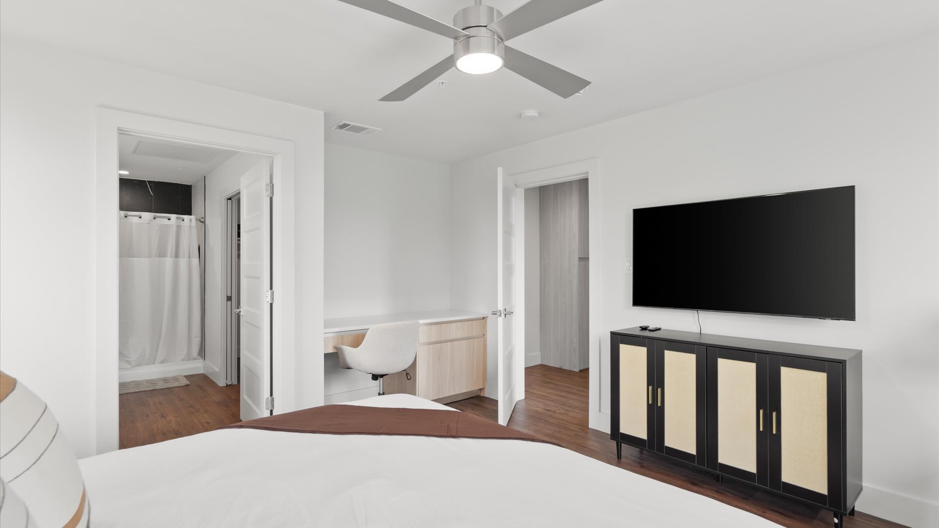 View of the bedroom showing a built-in desk with a white chair and light wood cabinetry, adjacent to an open doorway leading to a closet or another room. A large flat-screen TV is mounted on the wall opposite the bed.