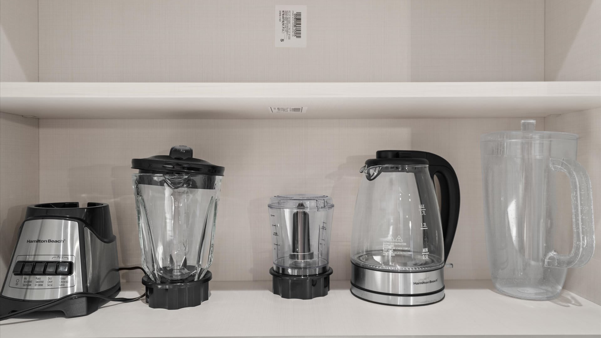 An organized kitchen cabinet shelf displaying various small appliances, including a Hamilton Beach blender, a smaller food processor, a Hamilton Beach electric kettle, and a clear plastic pitcher.
