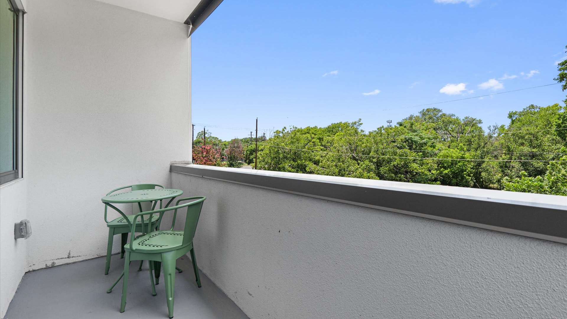 A small outdoor balcony features a green metal bistro table and two matching chairs, set against a white stucco wall with a gray railing, overlooking lush green trees and a clear blue sky.