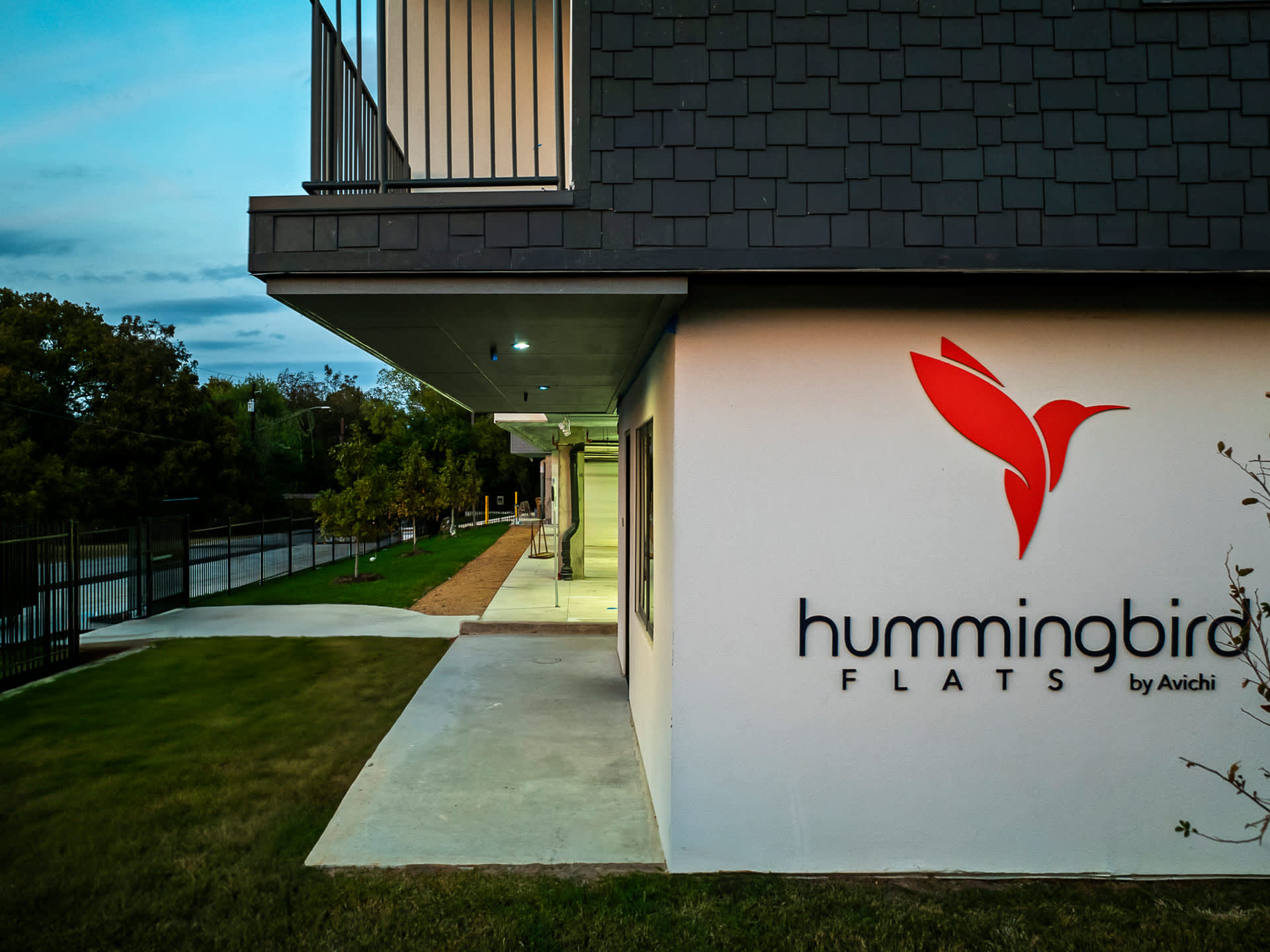The exterior of "Hummingbird Flats by Avichi" building features a white facade with a red hummingbird logo, dark gray siding, and a paved walkway leading to the entrance, surrounded by green grass and trees.
