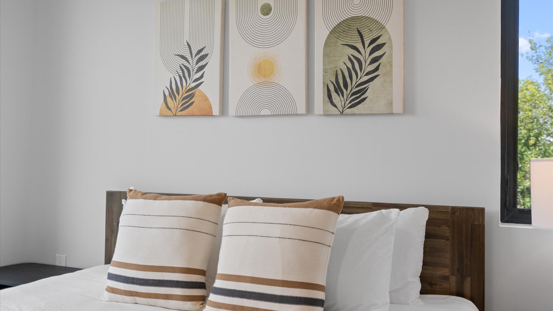 A bright bedroom features a wooden headboard with white and brown striped pillows, complemented by three abstract botanical art pieces on the white wall above. A window on the right offers a glimpse of greenery.
