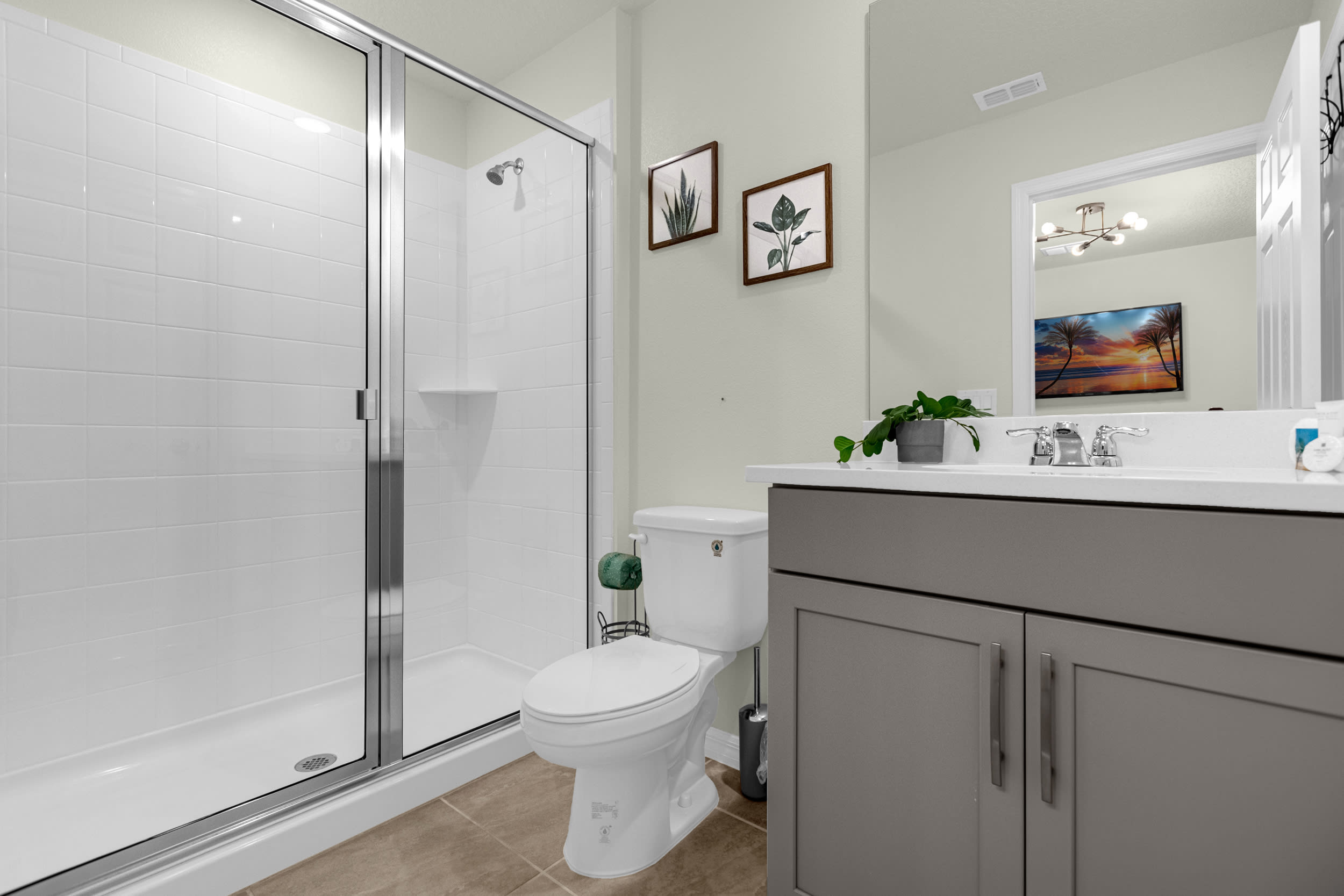 A clean, modern bathroom featuring a gray vanity with a white countertop and a large mirror, adjacent to a white toilet. It includes a walk-in shower with white tiled walls and a clear glass enclosure.