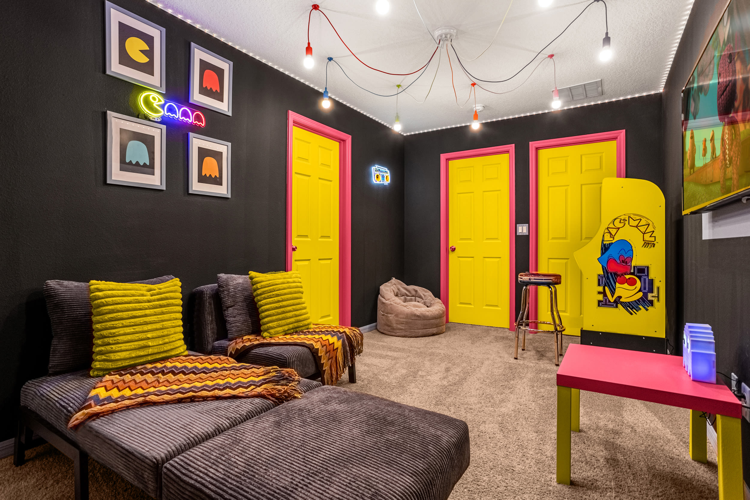 Playful game room with dark walls, bright yellow doors, and a gray sectional sofa with yellow accent pillows. Neon signs and framed Pac-Man art adorn the walls, creating a fun, retro atmosphere.